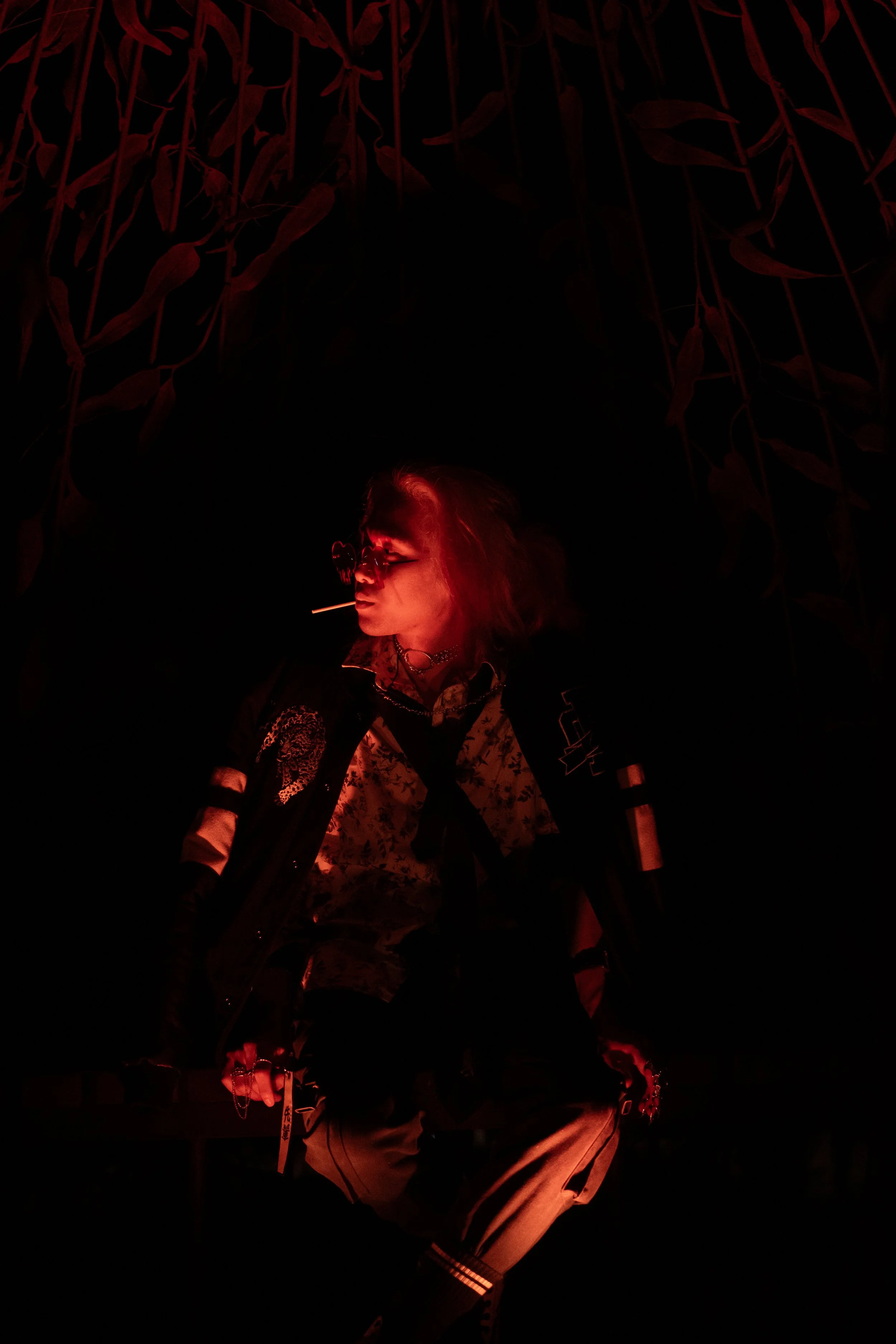 Person with long hair, glasses, wearing a jacket with patches, and holding a stick, illuminated by red lighting, with a dark background containing hanging plant leaves.