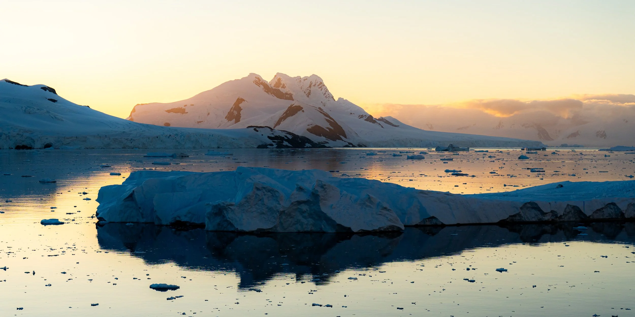 Snow-capped mountains at sunset with icebergs floating in calm water.