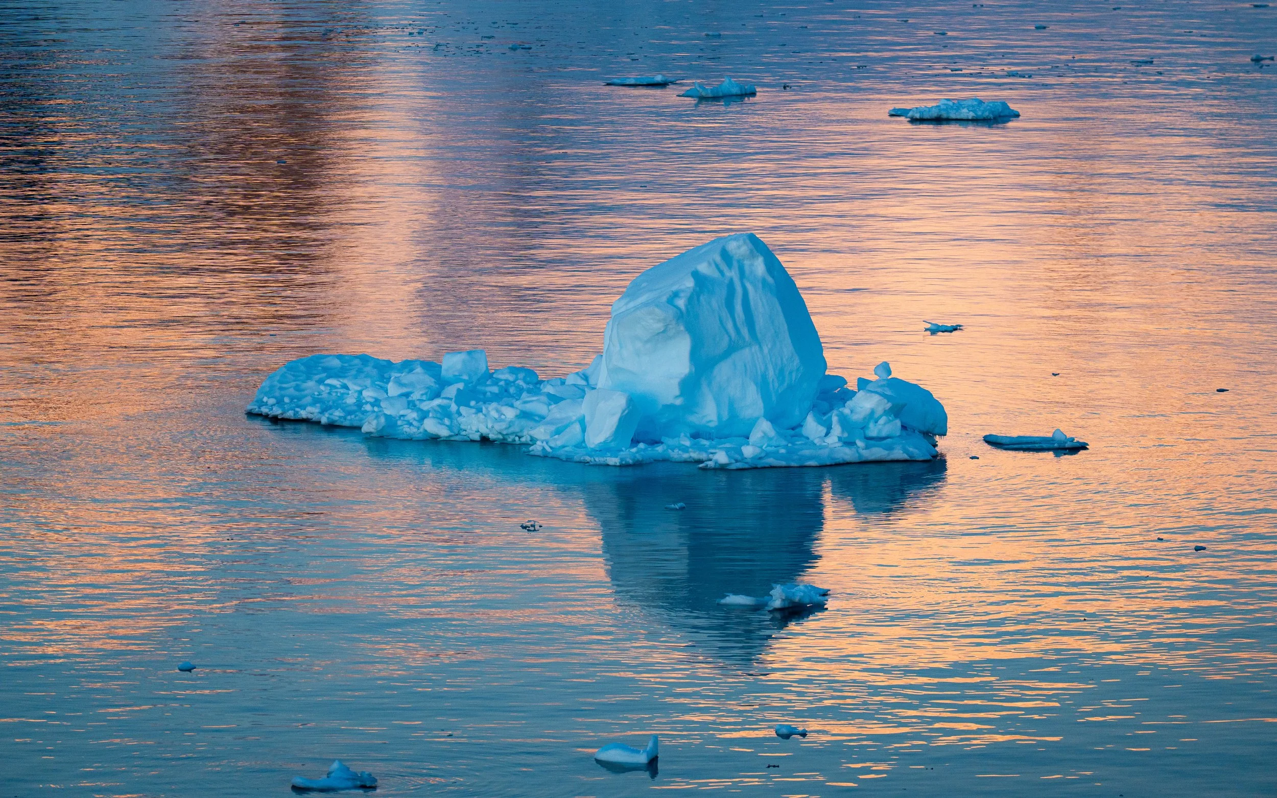Iceberg floating in the ocean during sunset with smaller ice chunks around it.