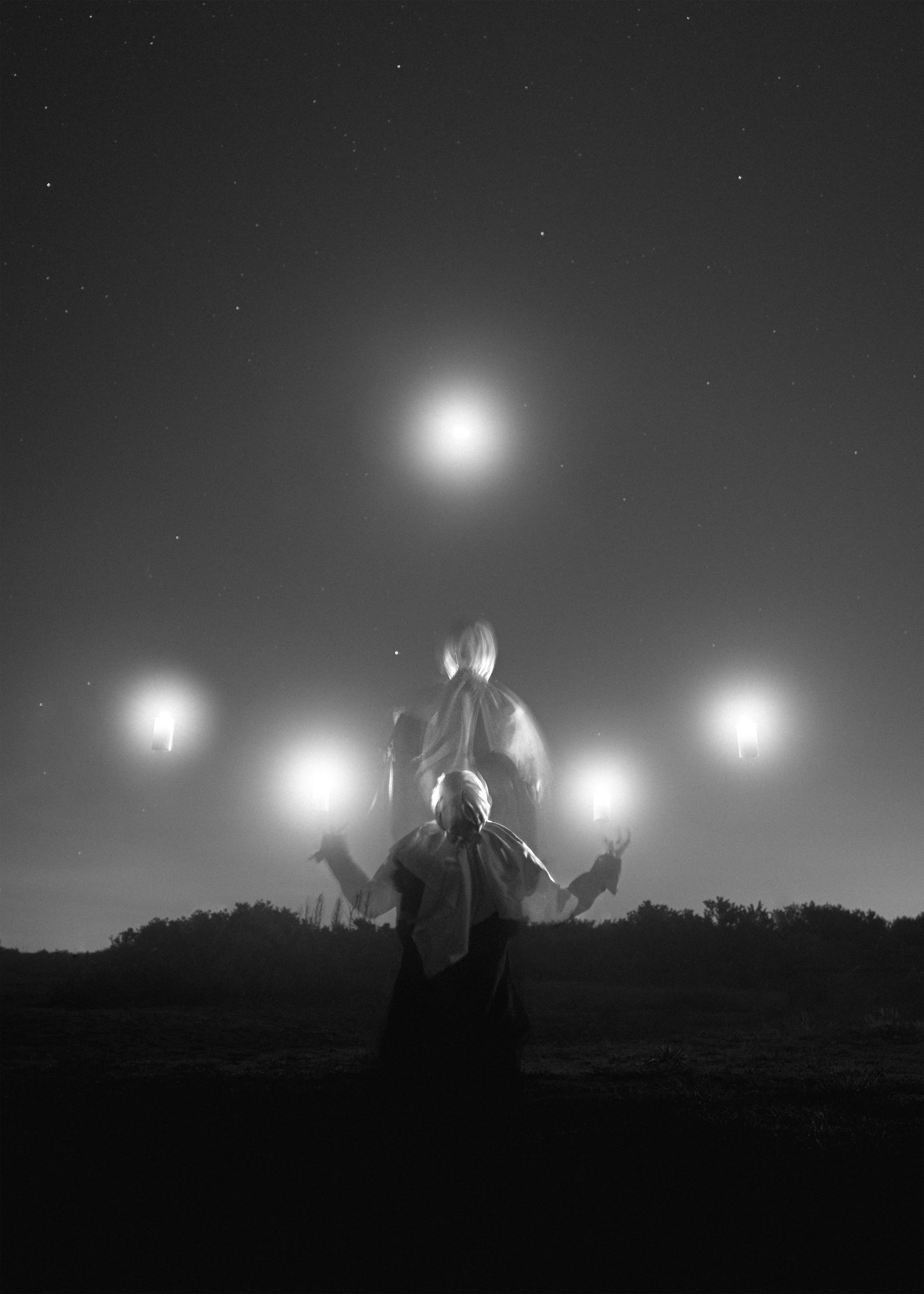 Person with long hair and traditional clothing performing a fire dance outdoors at night, with a clear sky and stars above, and flames and lanterns illuminating the scene.