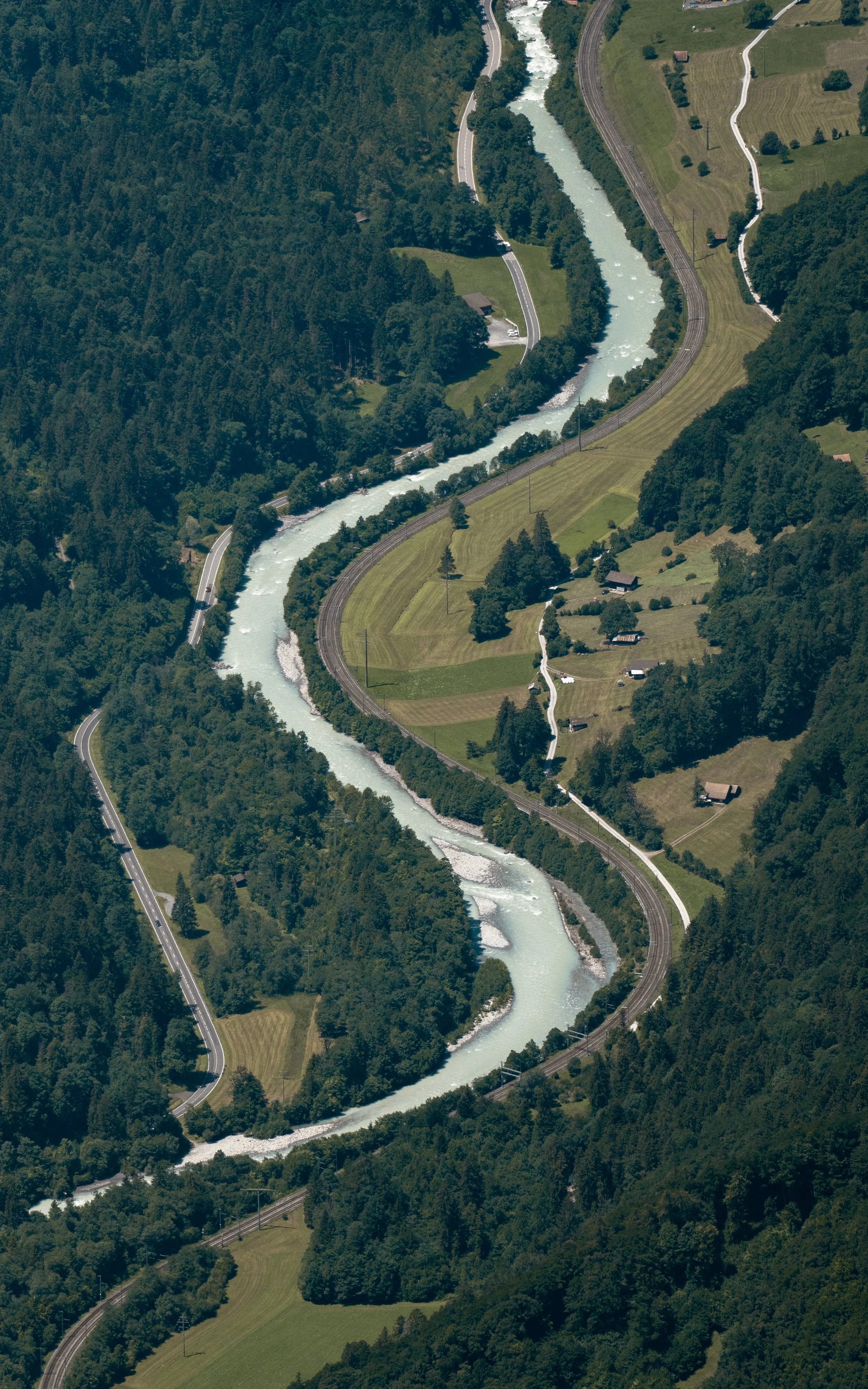 Aerial view of a winding river through a forested, hilly landscape with train tracks and small houses nearby.