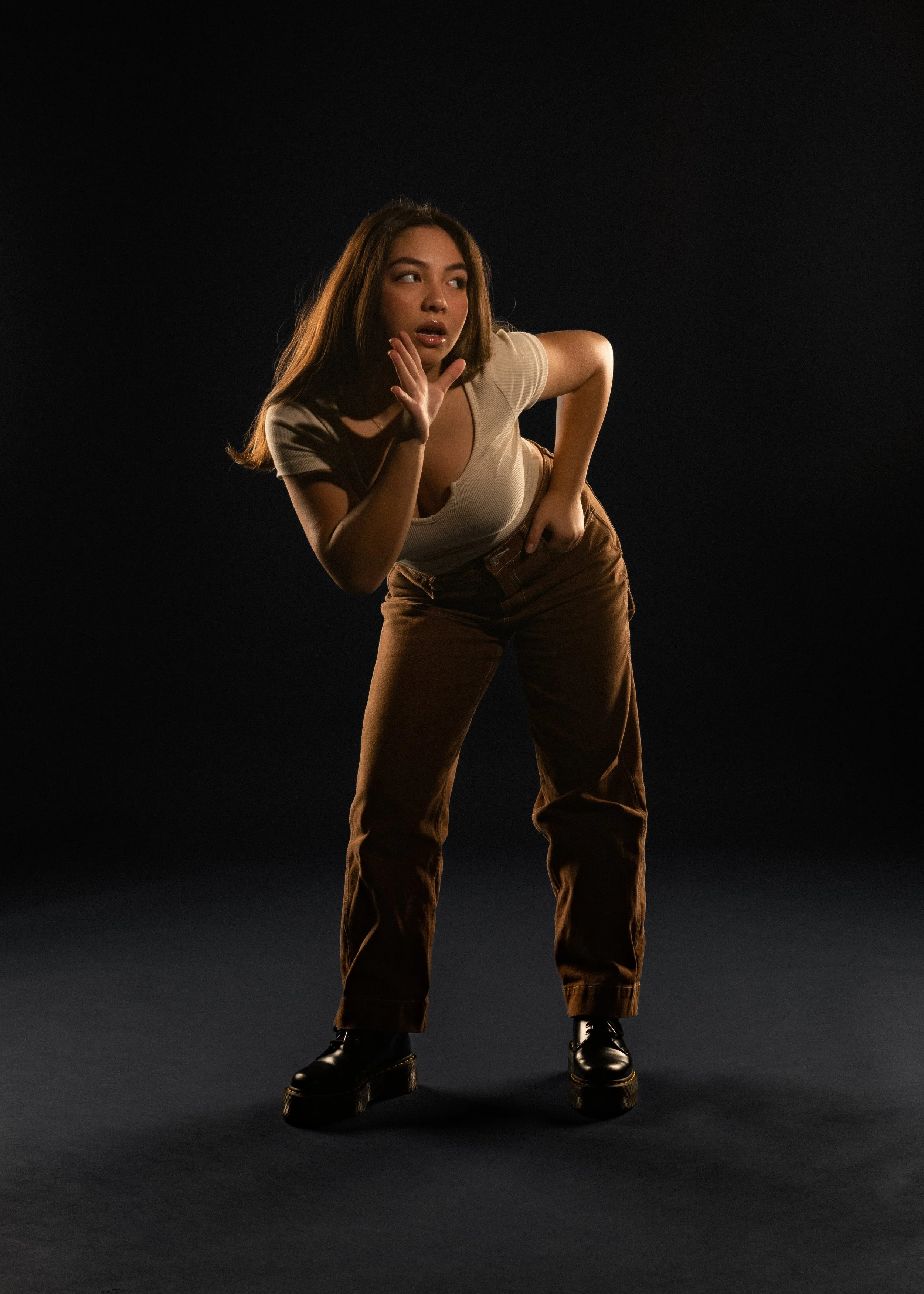 A woman with long brown hair wearing a beige t-shirt, brown pants, and black boots standing against a black background, striking a pose with one hand near her face.