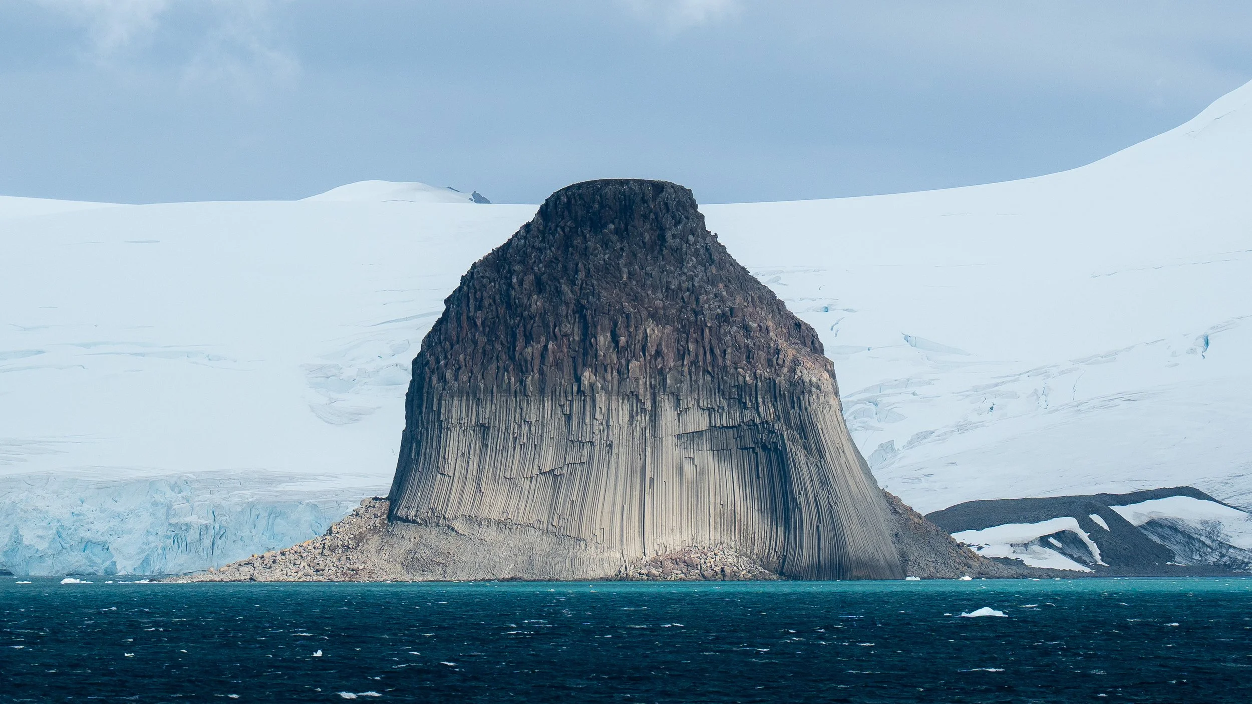 A large dark mountain isolated in icy waters, with glacial ice and snow-covered land in the background under a cloudy sky.