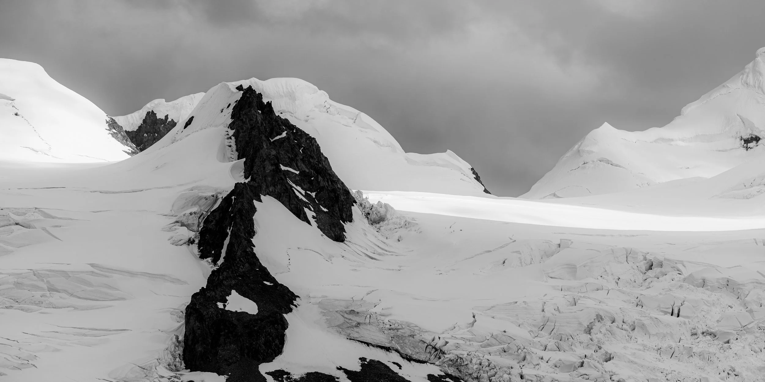 Black and white photo of snowy mountains and glaciers with dark rocky peaks, overcast sky.