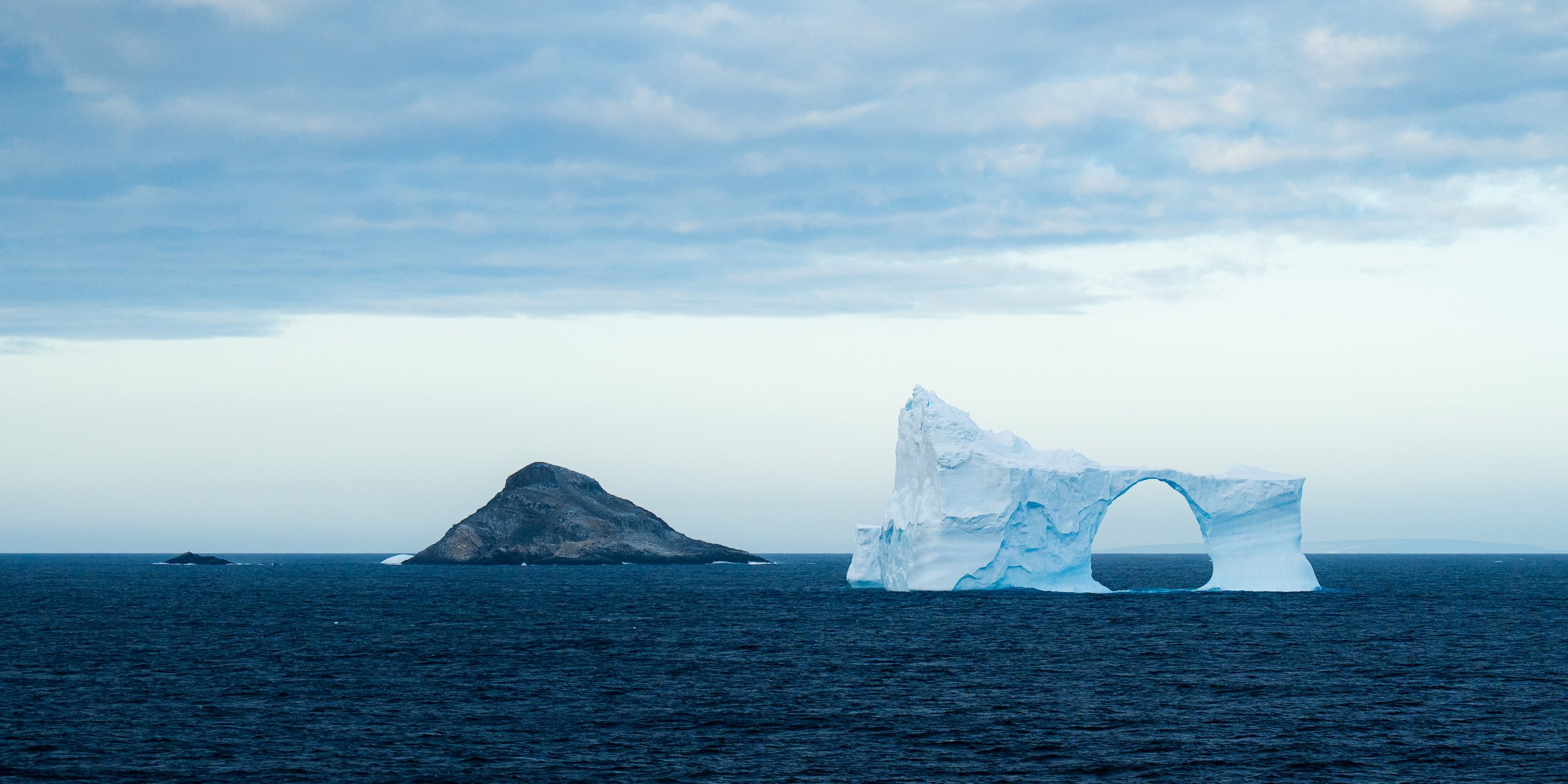 A large iceberg with a natural arch formation floating in the ocean near a rocky island seen under a cloudy sky.