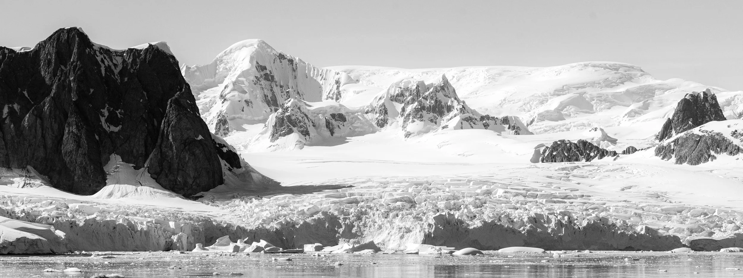 Black and white photo of snow-covered mountains with rugged dark rocks in the foreground and icy water at the bottom.