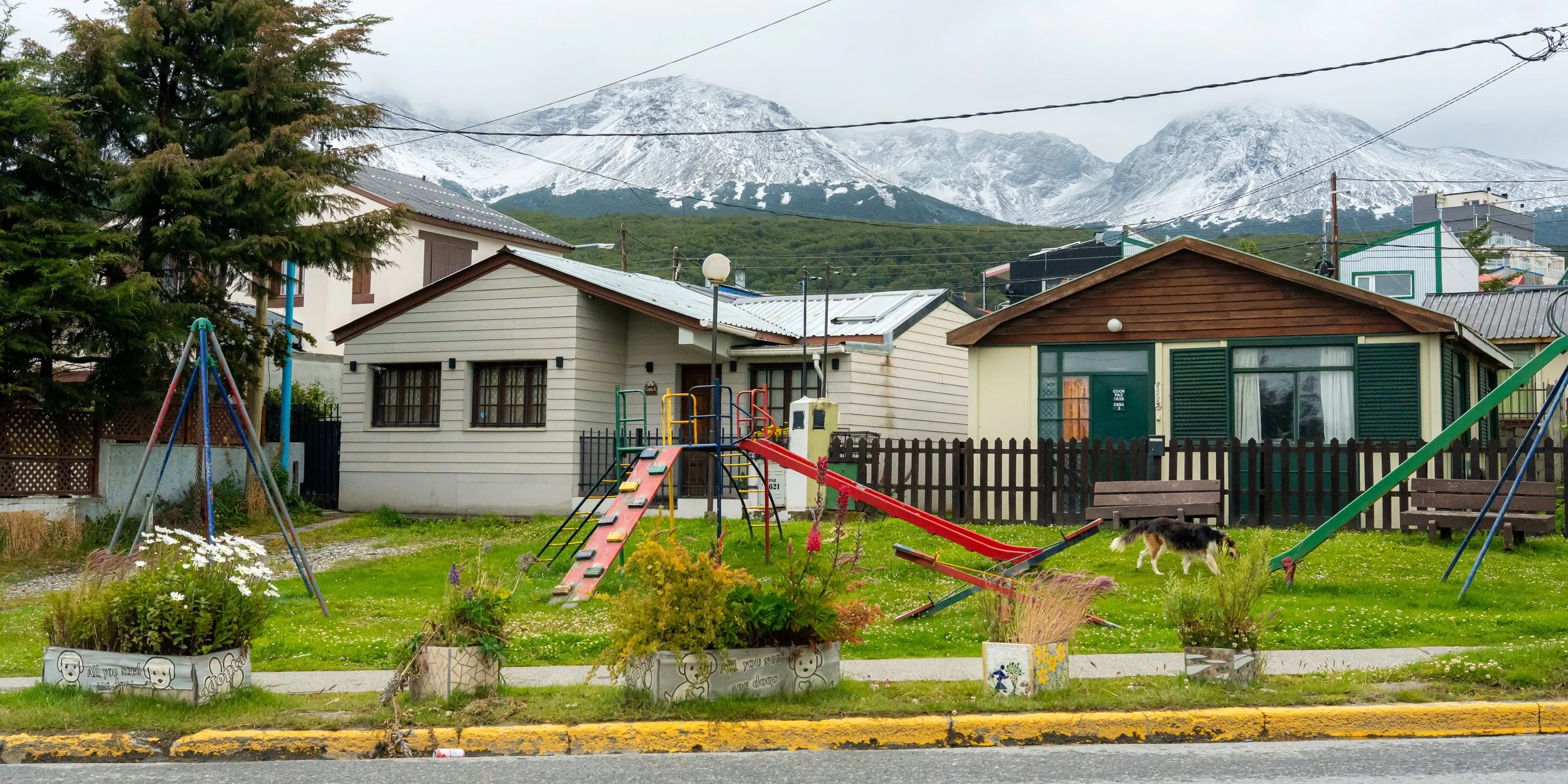 A suburban backyard with playground equipment, benches, potted plants, and houses under snow-capped mountains in the background.