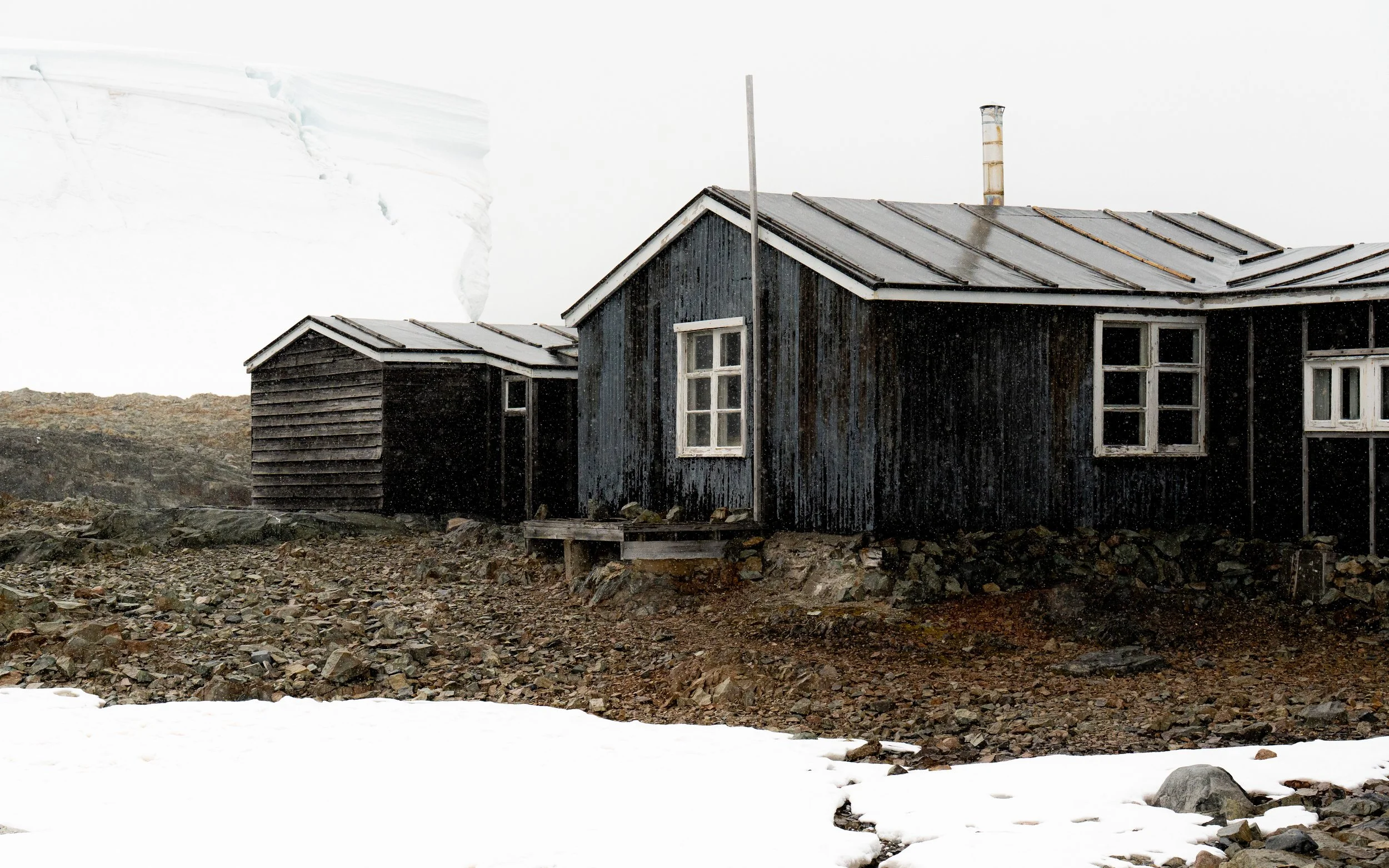 A black wooden house with white window frames located on rocky terrain, with snow in the foreground and an iceberg or glacier in the background.