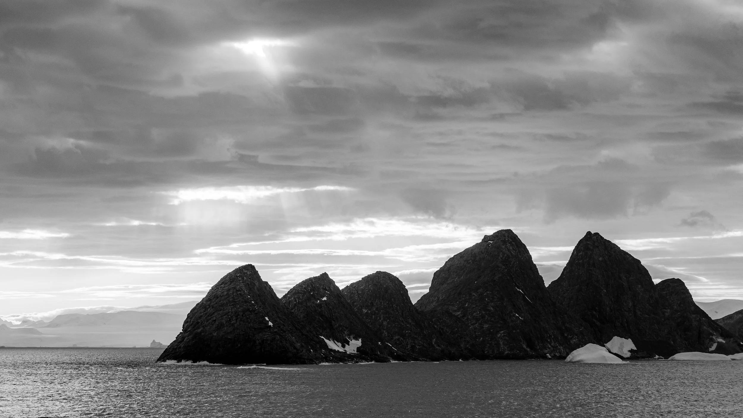 Black and white photo of rugged mountain peaks near a body of water, with dramatic cloud-filled sky and sunlight breaking through.