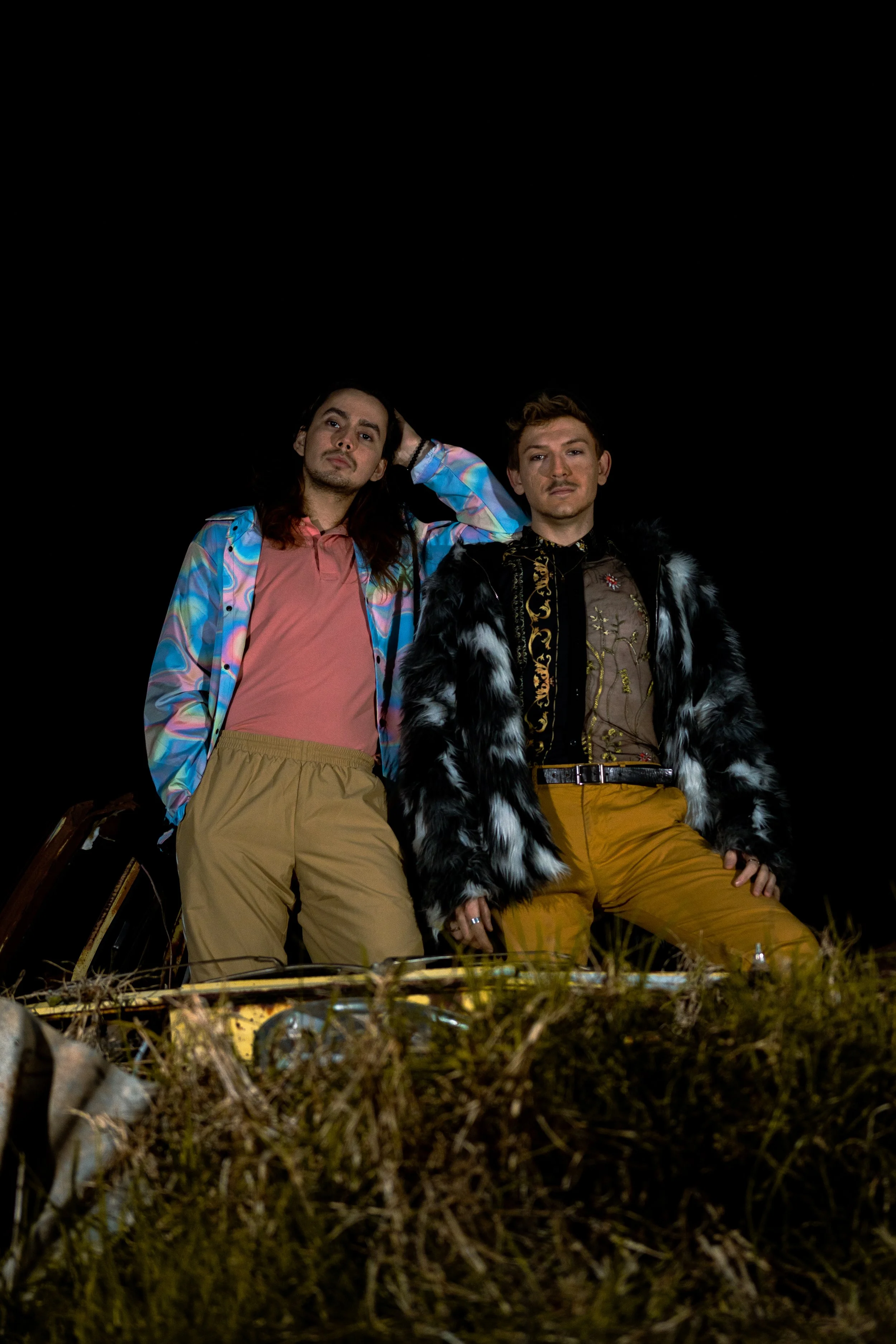 Two men standing outdoors at night, dressed in colorful, eclectic outfits. One wears a patterned jacket and tan pants, the other a fur-lined coat with gold embroidery and mustard-colored pants. They are surrounded by dry grass and debris.
