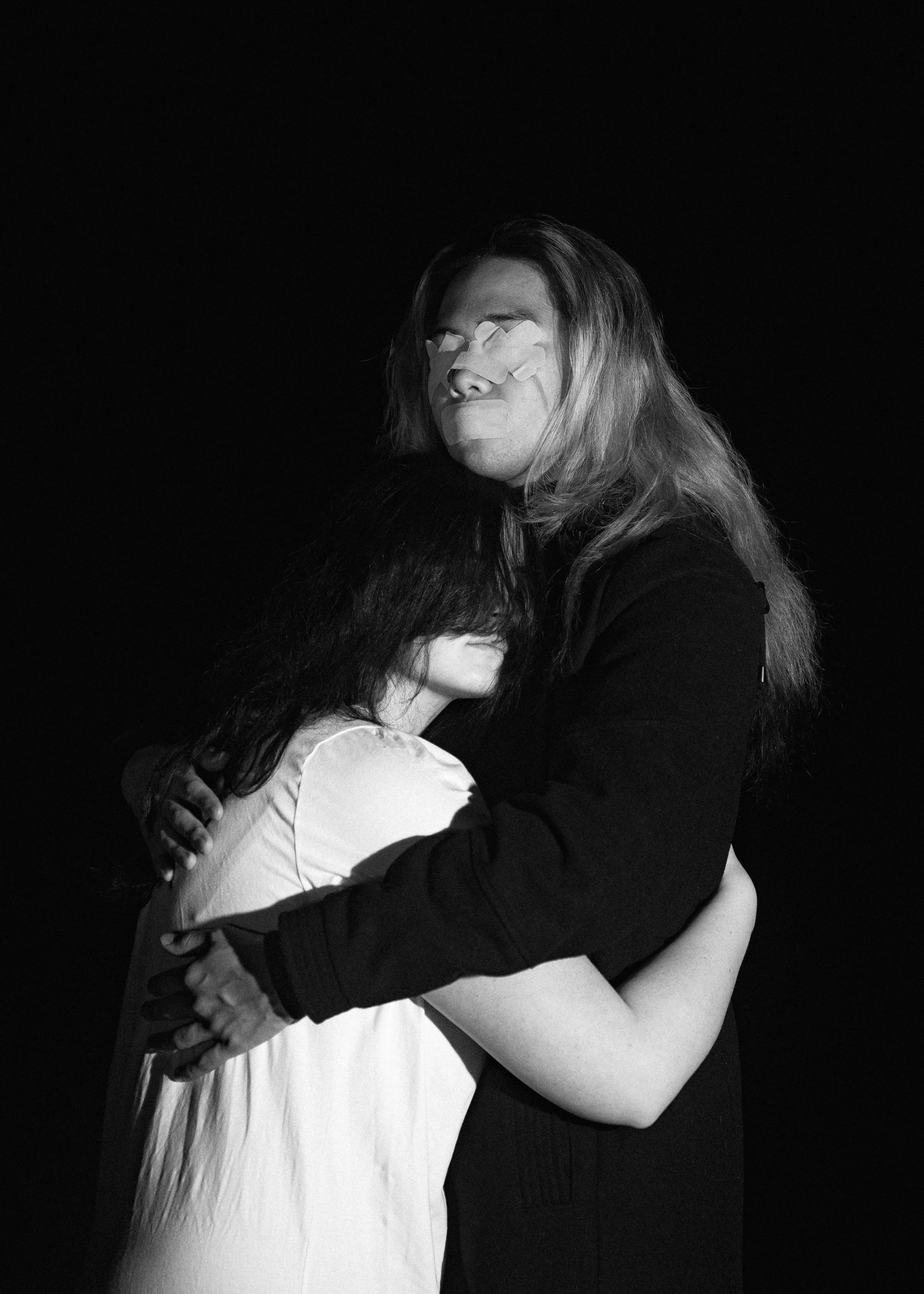 Two women hugging tightly, one with long hair and the other with short dark hair, against a black background.