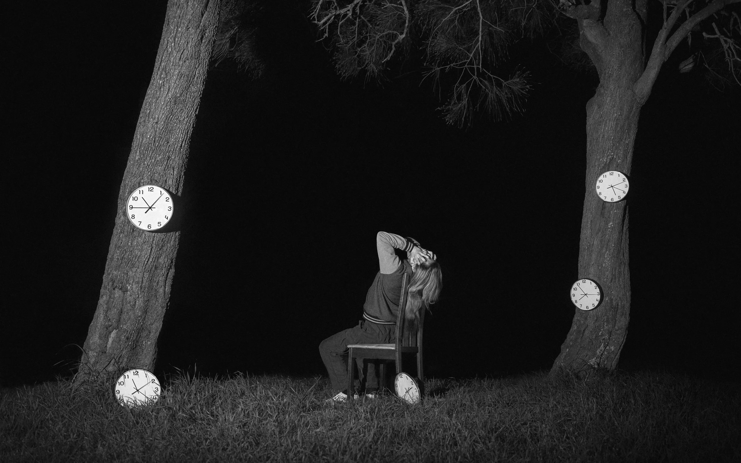 A person with long hair and a jacket sitting on a chair with their head leaning back, surrounded by trees and clocks hanging on the trees and on the ground, at night.