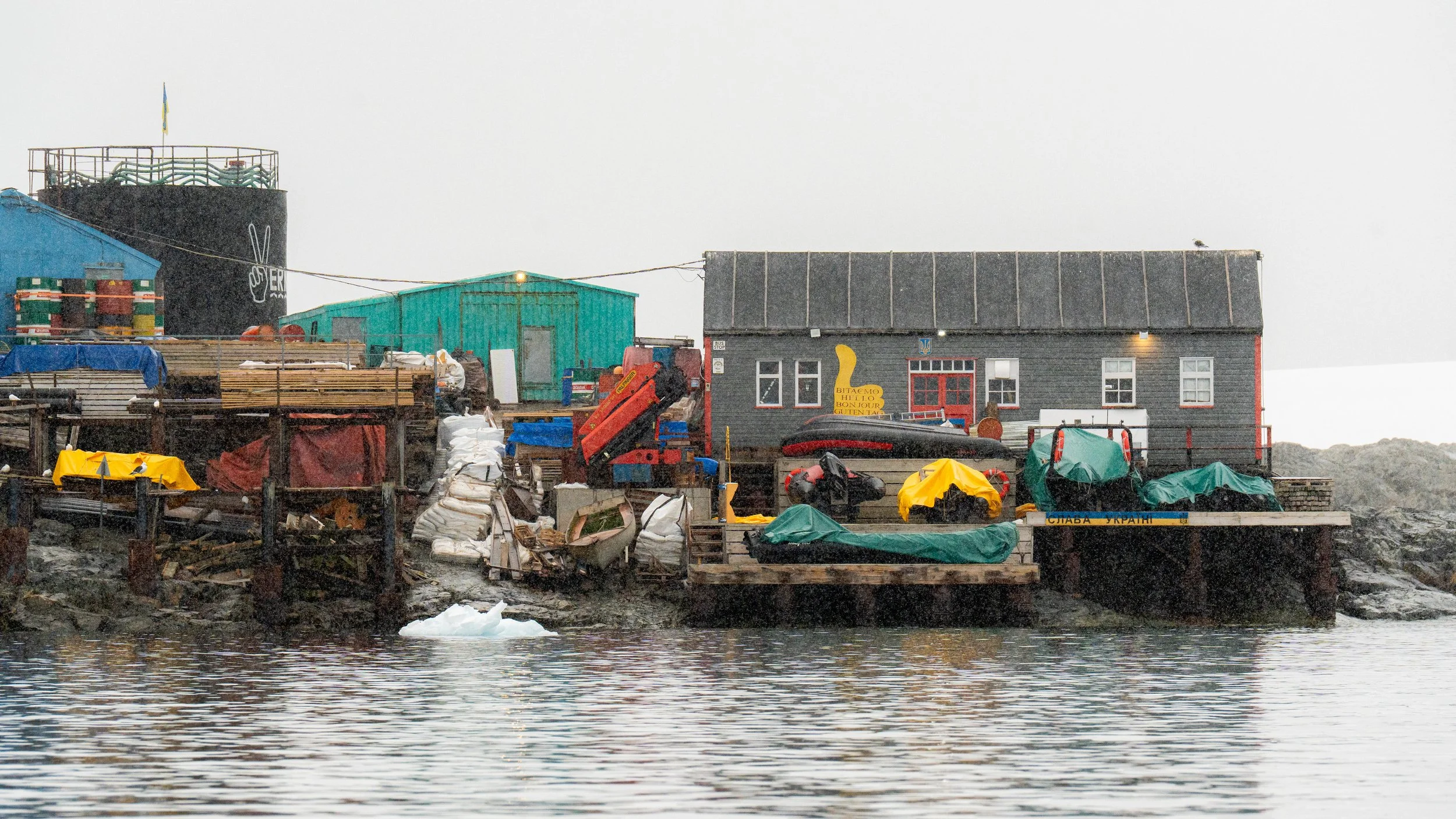 A dockside scene with houses and structures built on stilts over the water, including a gray house with red trim, a teal shed, a large black tank, and various boats and equipment, amid a gray, overcast sky.