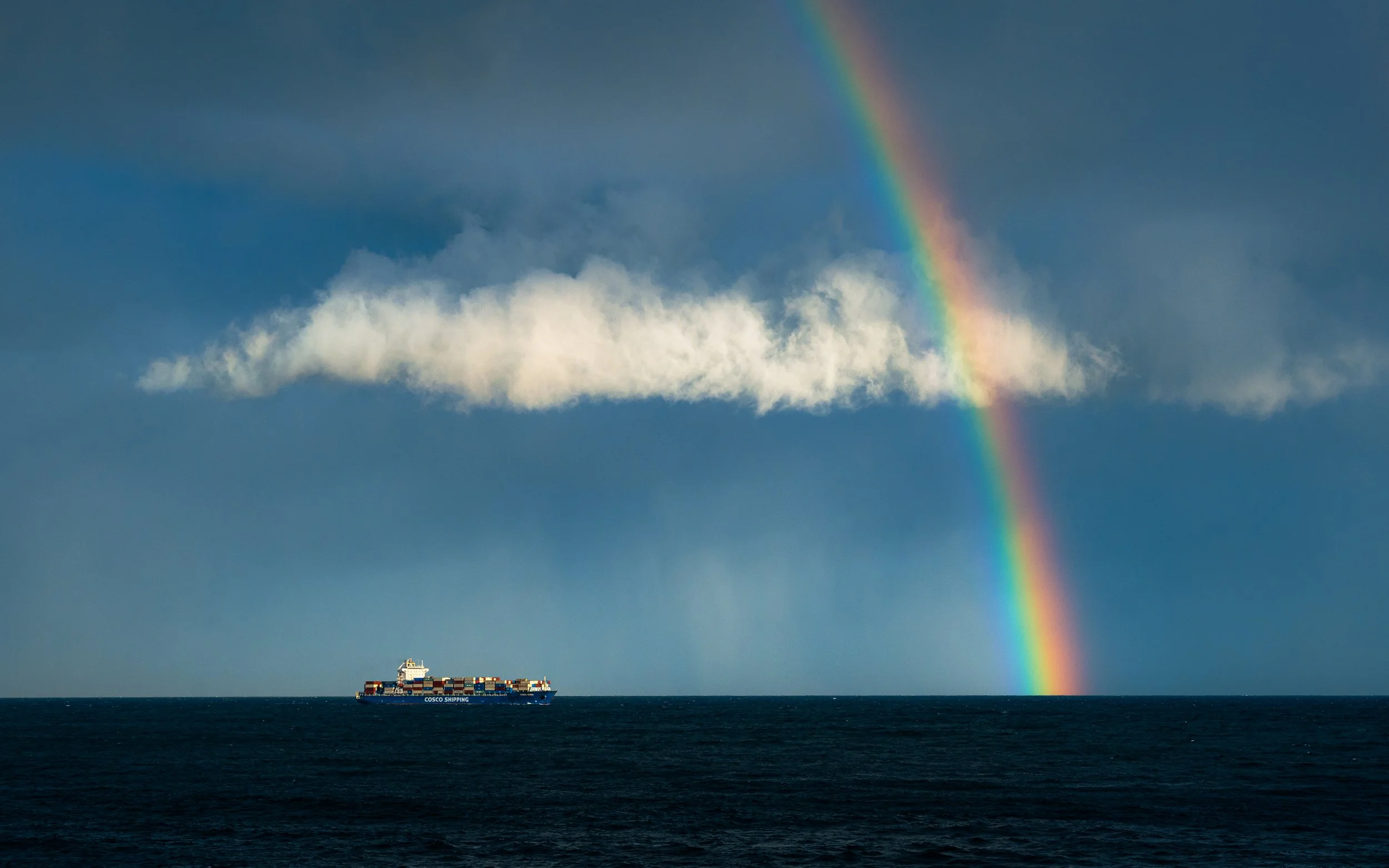 A cargo ship labeled 'Cosco Shipping' sailing on the ocean with a rainbow arching across a blue sky with scattered clouds.