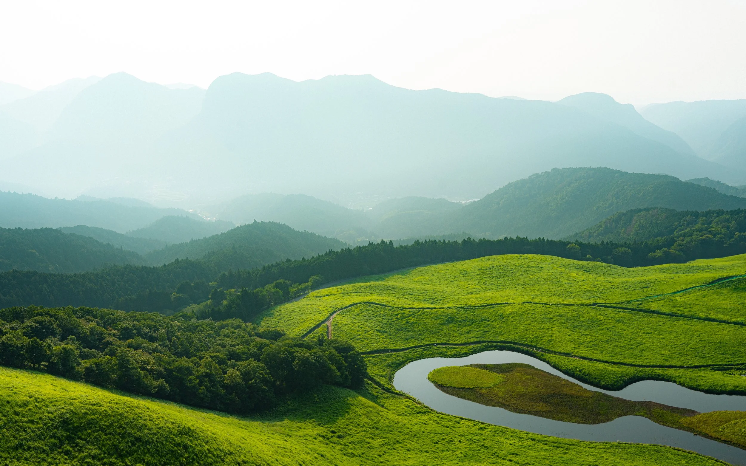Green rolling hills surrounding a winding river, with distant mountainous landscape under a hazy sky.