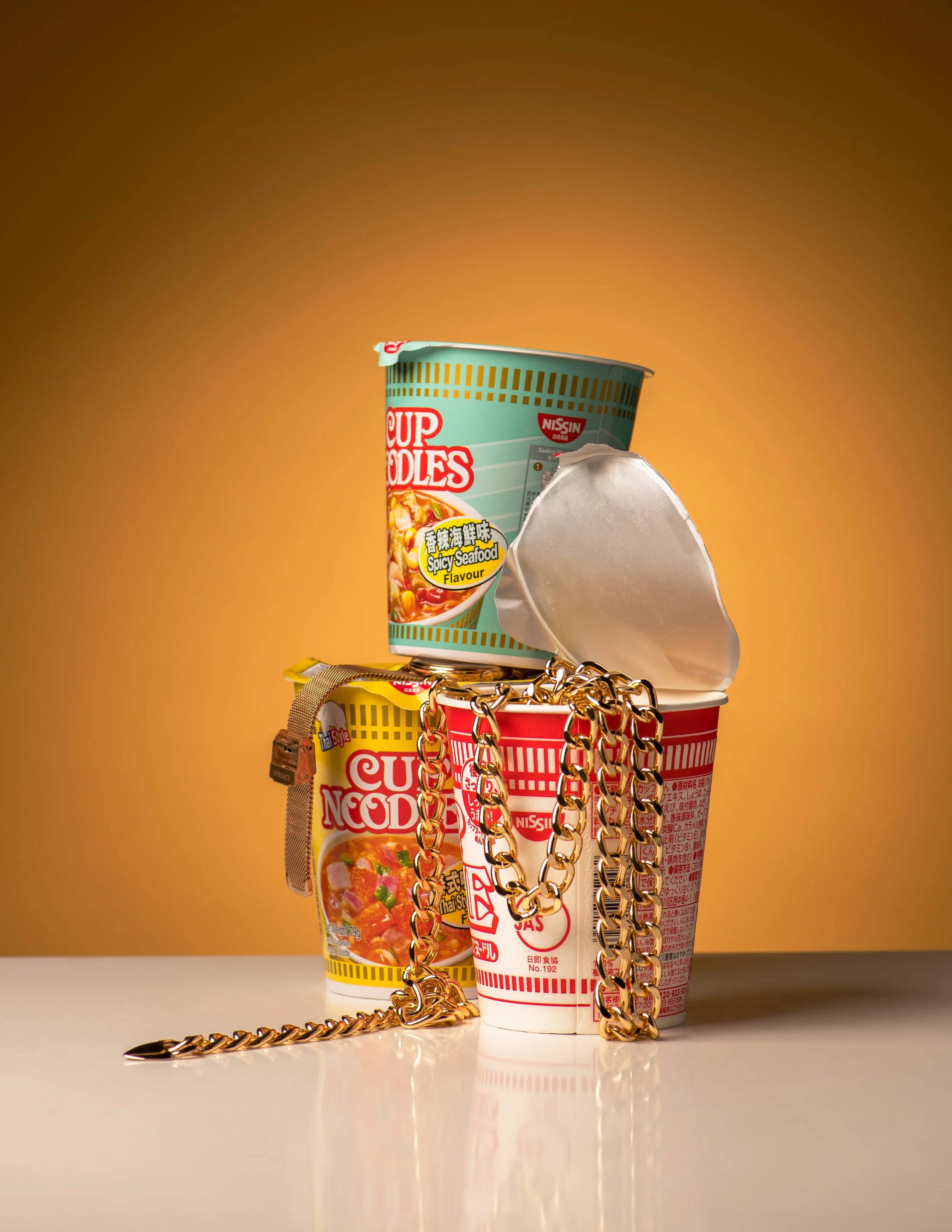 A collection of instant noodle containers, including a green Nissin cup noodle with spicy seafood flavor and a red and white cup noodle container, all stacked with gold chains and jewelry draped over them, on a white surface with a yellow background.