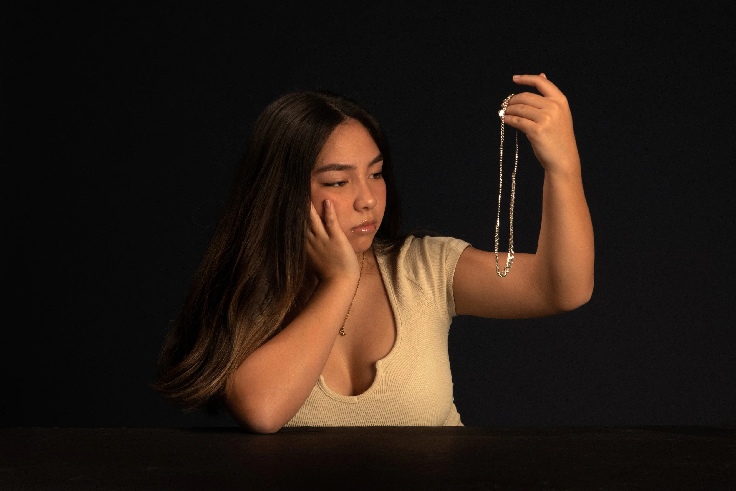 Young woman with long brown hair, looking thoughtful, holding a silver necklace in her right hand, and resting her chin on her left hand, against a dark background.