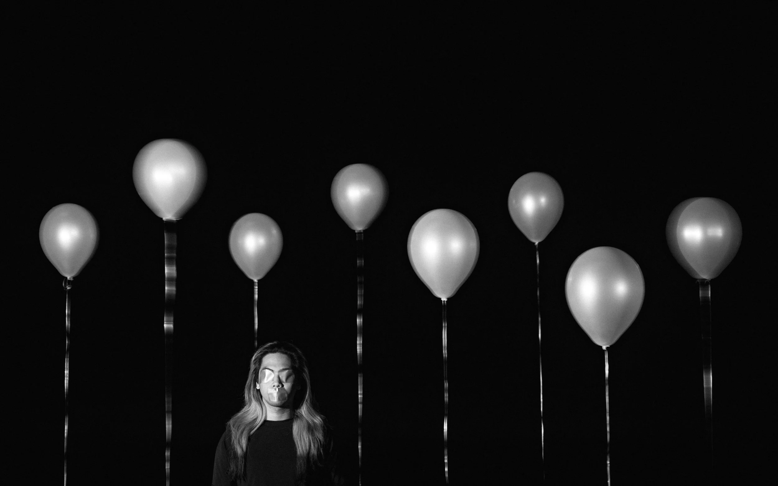 Black and white photo of a woman with long hair standing in front of balloons, with the top of her head and part of her face visible, and the balloons floating above her against a dark background.