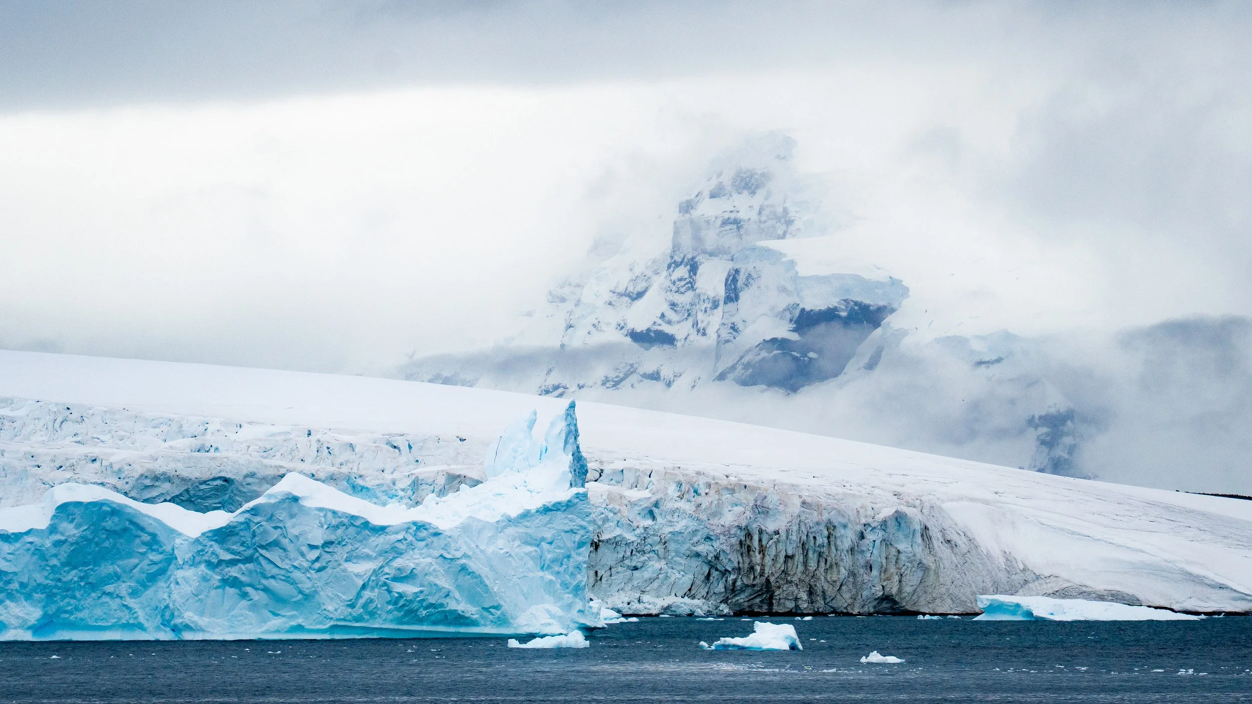 Iceberg floating in ocean with snow-covered mountain in background, partly obscured by clouds.