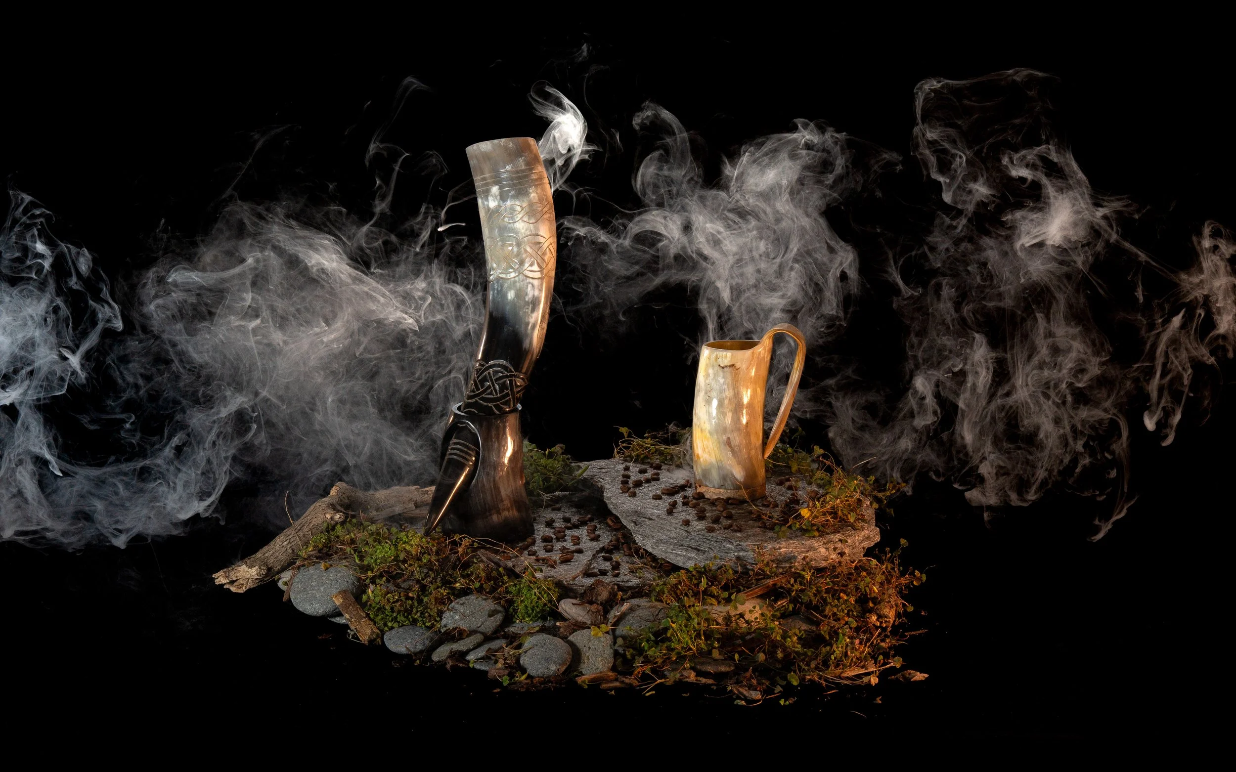 Two carved horns, one with smoke rising, placed on a wooden surface with small stones, greenery, and scattered pebbles, against a black background.