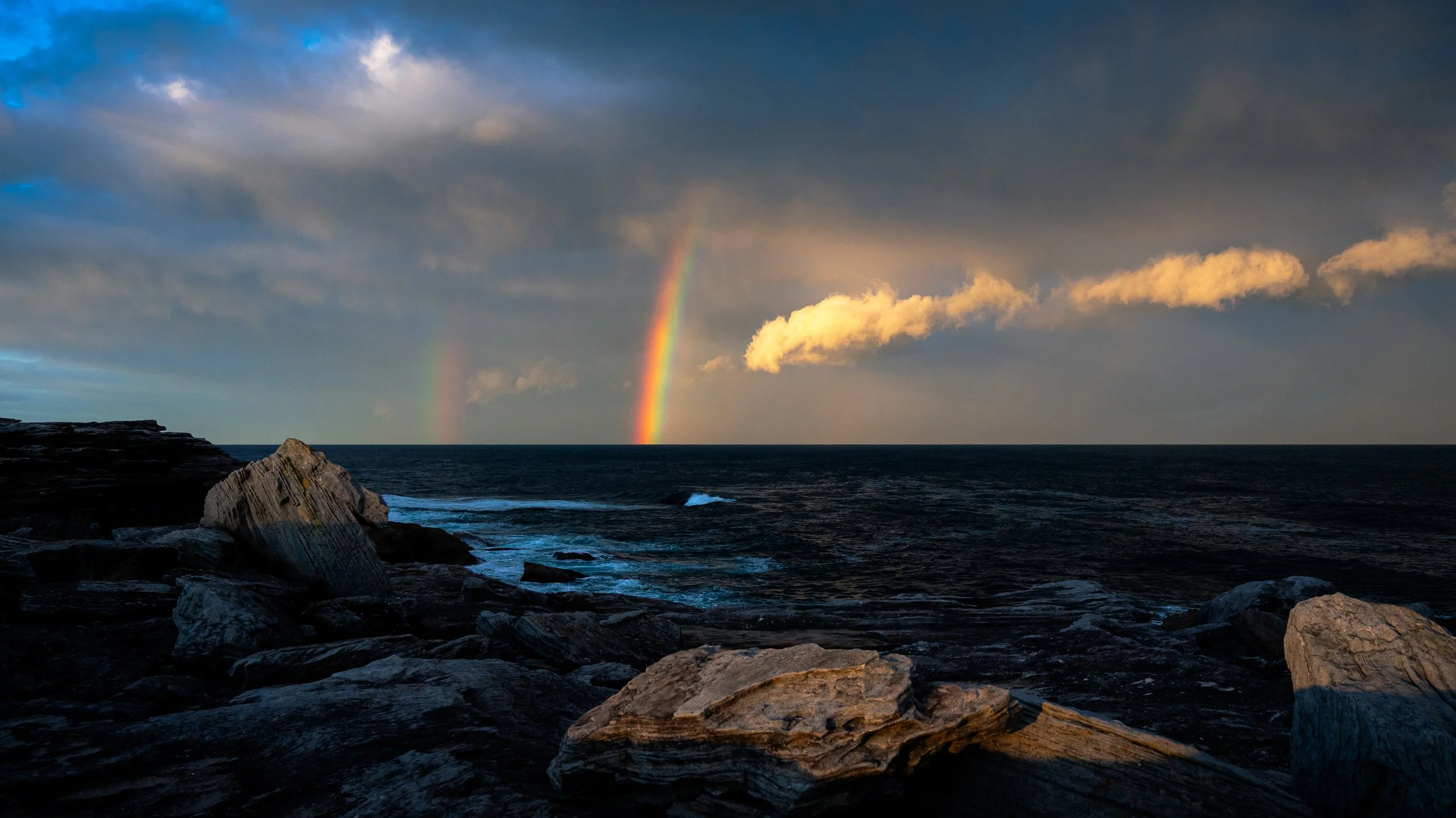 View of the ocean with rocky shoreline in the foreground, dark clouds in the sky, and two rainbows visible in the distance.