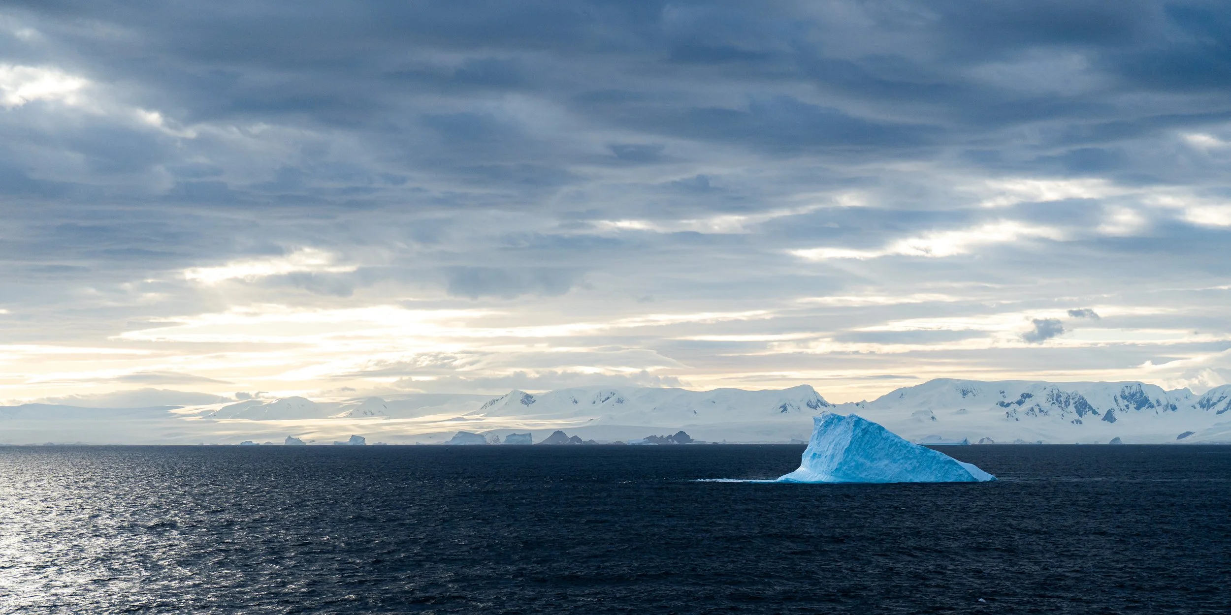A vast ocean with a large blue iceberg in the foreground and snow-covered mountains in the background under a cloudy sky.