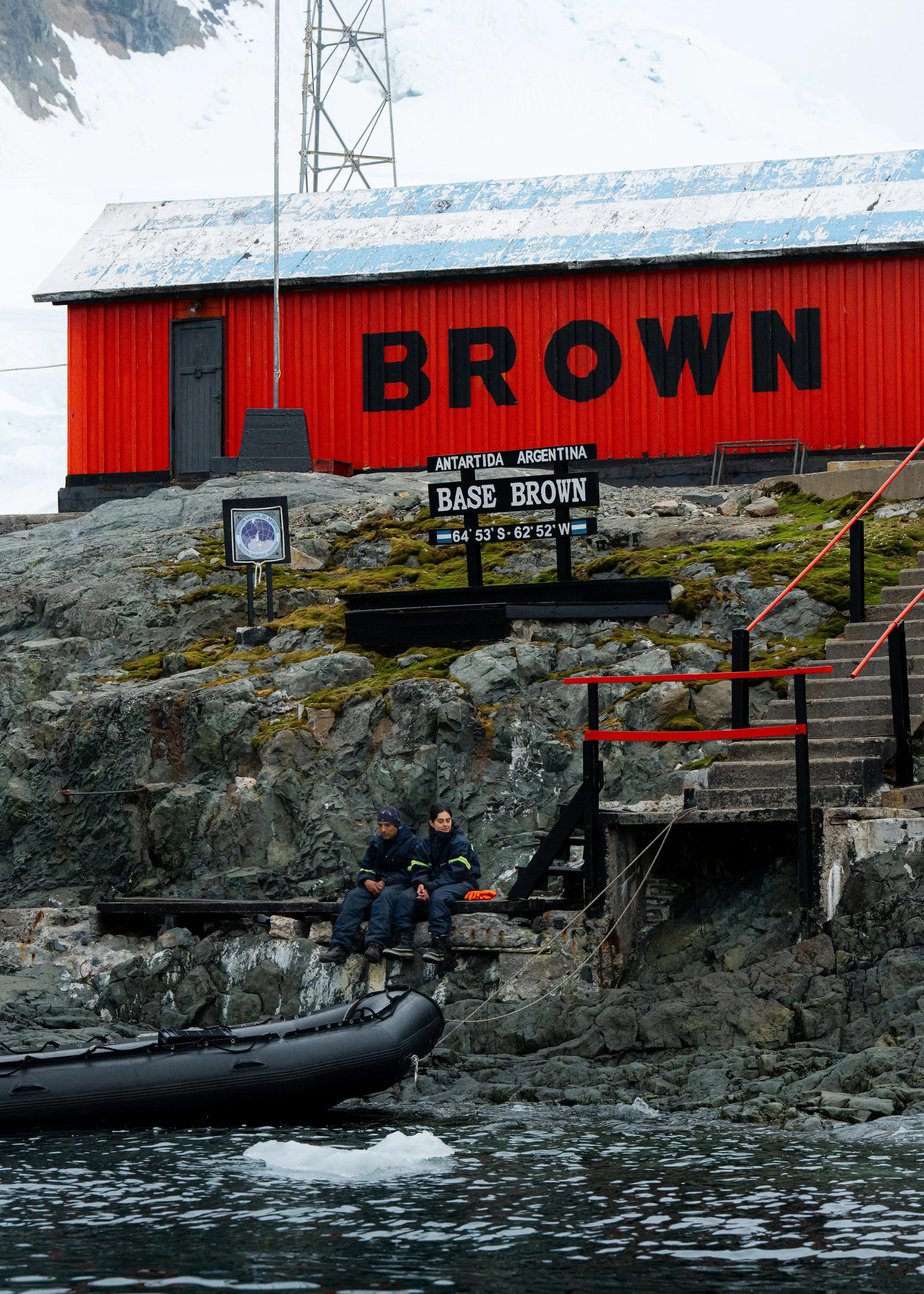 Red building with large black letters spelling 'BROWN' on the side, situated on rocky terrain near water; sign indicating location as Antarctica, Base Brown, with coordinates 64°53'S 62°52'W; people sitting on rocks near water, inflatable boat at wat