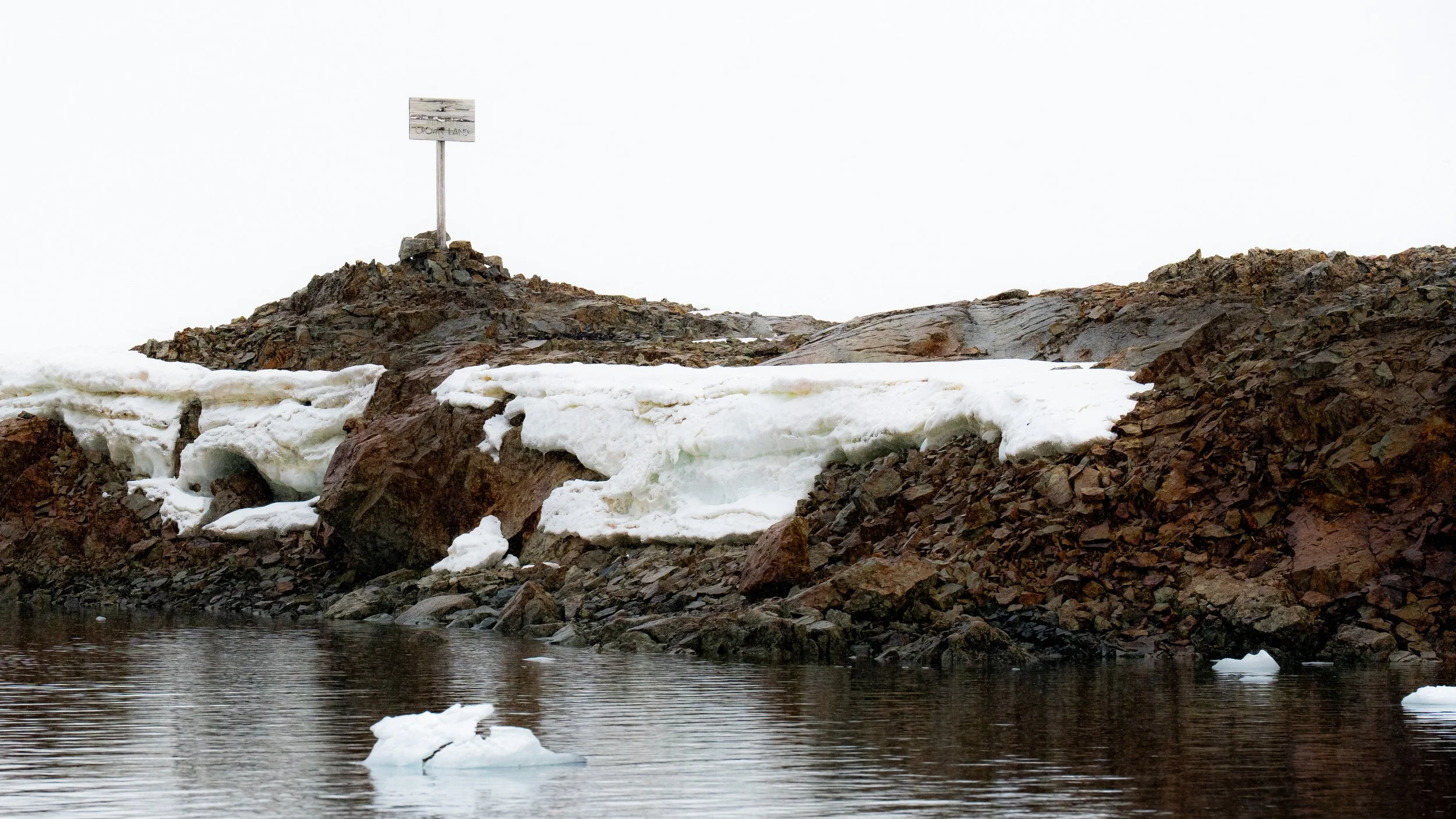 A rocky shoreline with ice and snow along the edge of a body of water, with a sign on a pole on top of the rocks.