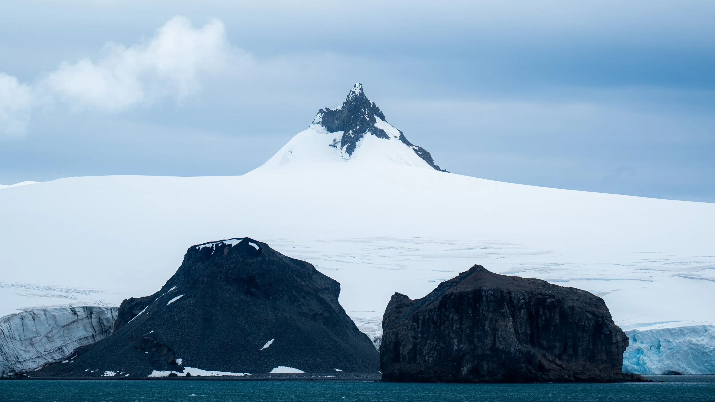 Mountain with snowy peak and glaciers surrounded by dark rocks in a cold landscape, with a turquoise body of water in the foreground and a cloudy sky above.