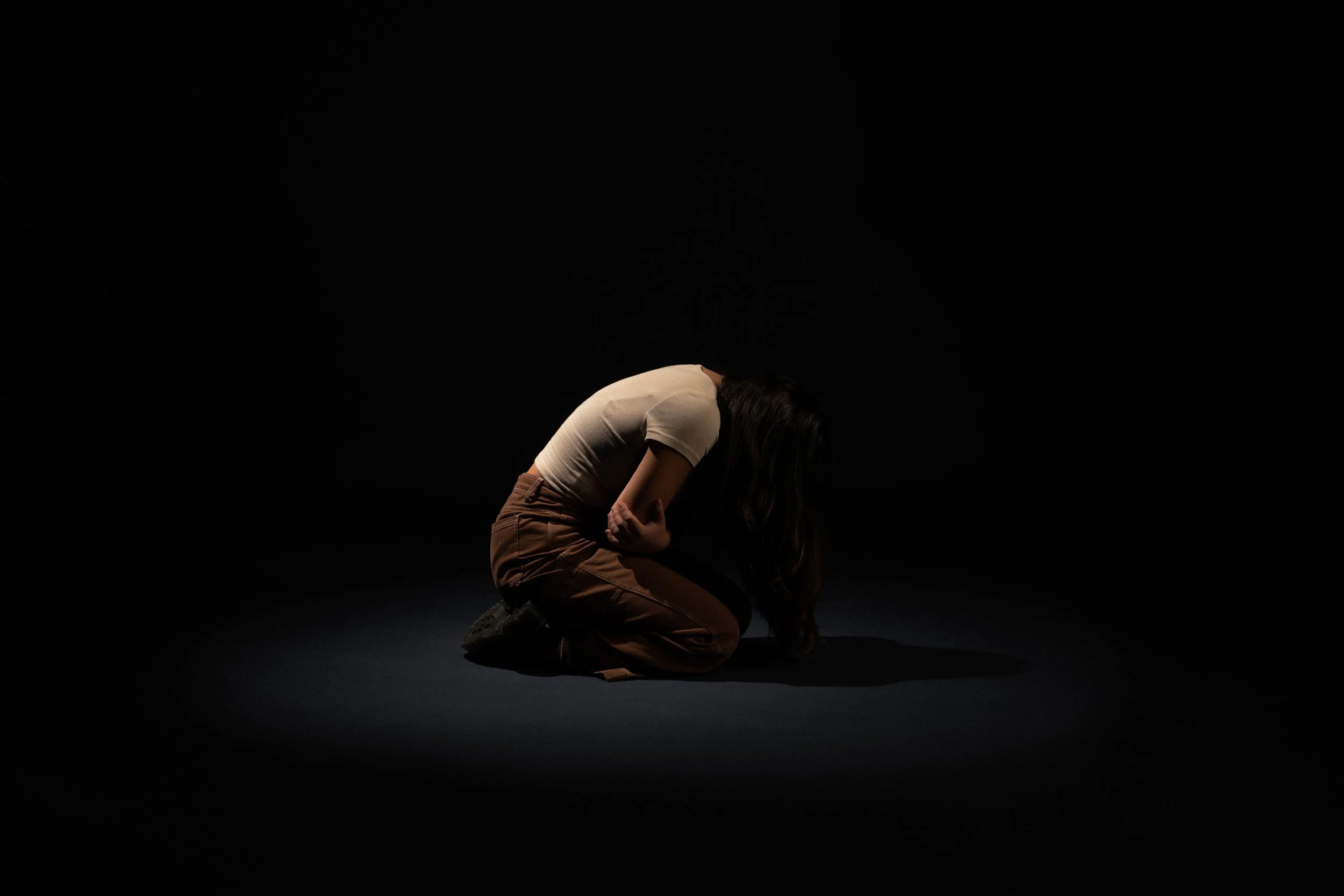 A person kneeling on the floor with their head bowed and hands clasped, in a dark setting with a black background.