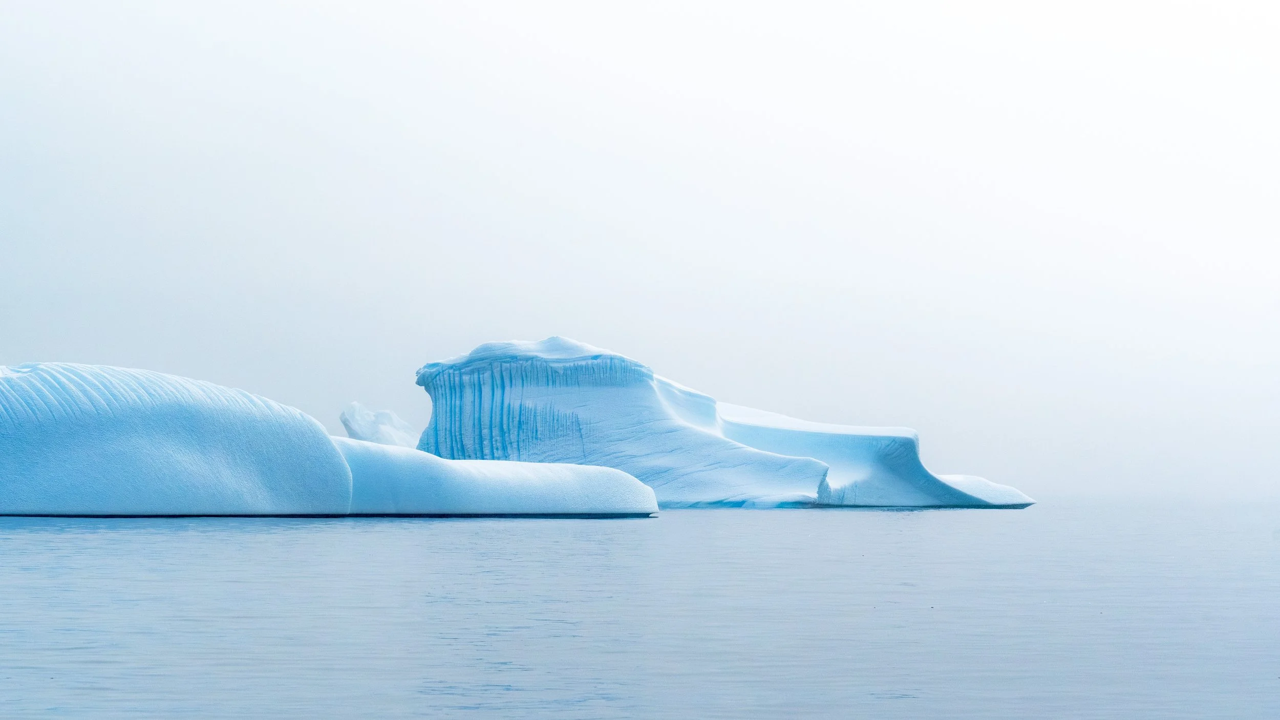 Large iceberg floating in cold, calm water with a clear, pale sky in the background.