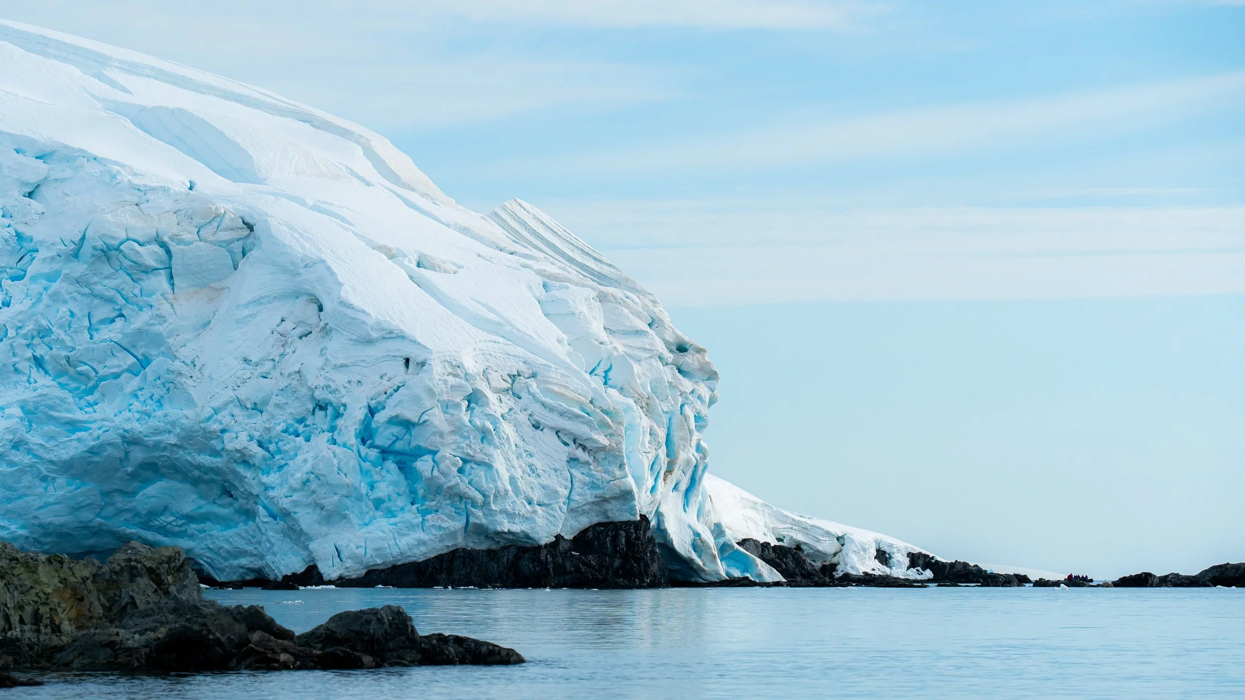 A large glacier with blue and white ice extending into a calm body of water, with rocky terrain in the foreground and a clear sky in the background.