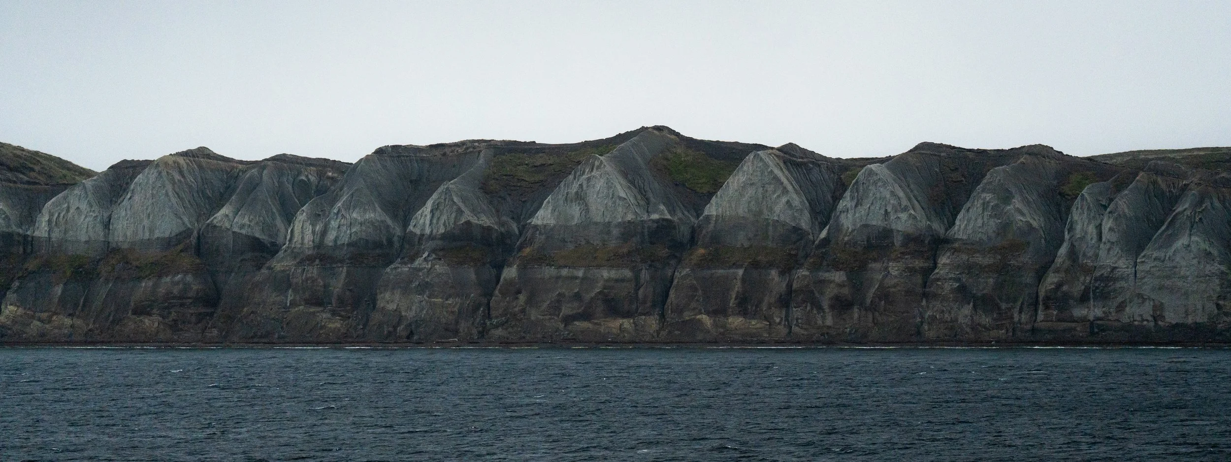 Cliffs with steep, gray rocky faces overlooking dark water under an overcast sky.