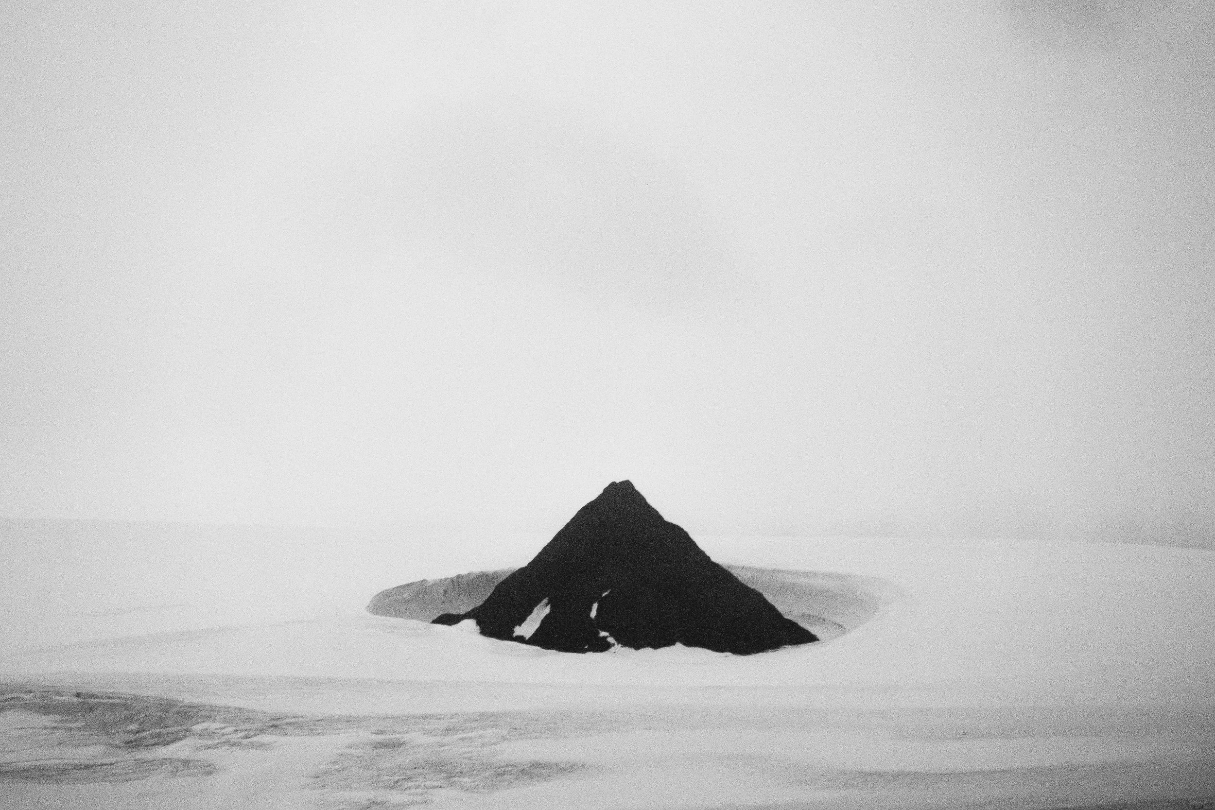 Black and white photo of a mountain landscape with a dark mountain peak in the foreground and a distant mountain range in the background, under a cloudy sky.