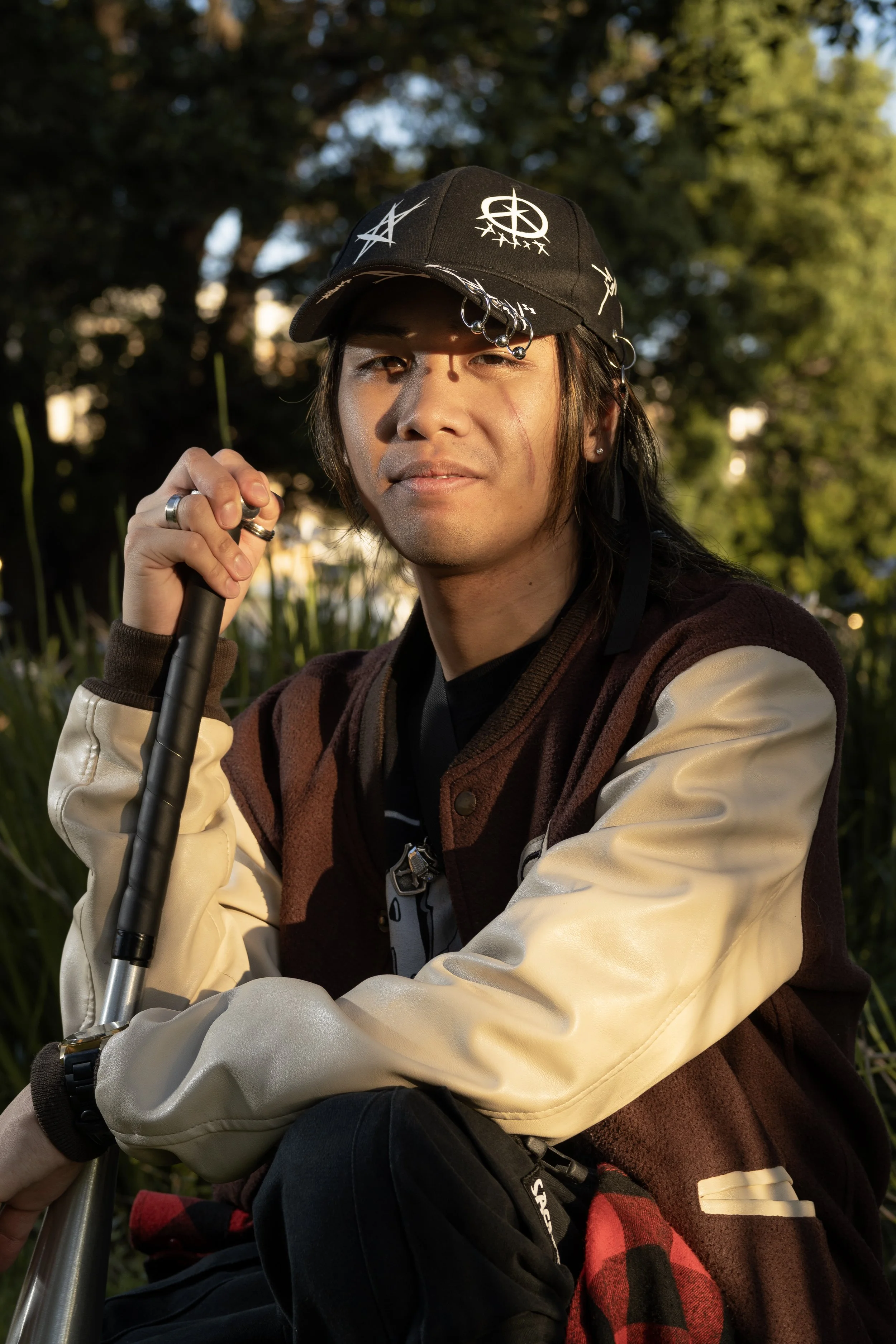 Young man with long dark hair and piercings, wearing a graphic black cap with metal rings and symbols, sitting outdoors during golden hour with green trees in the background, holding a walking stick.