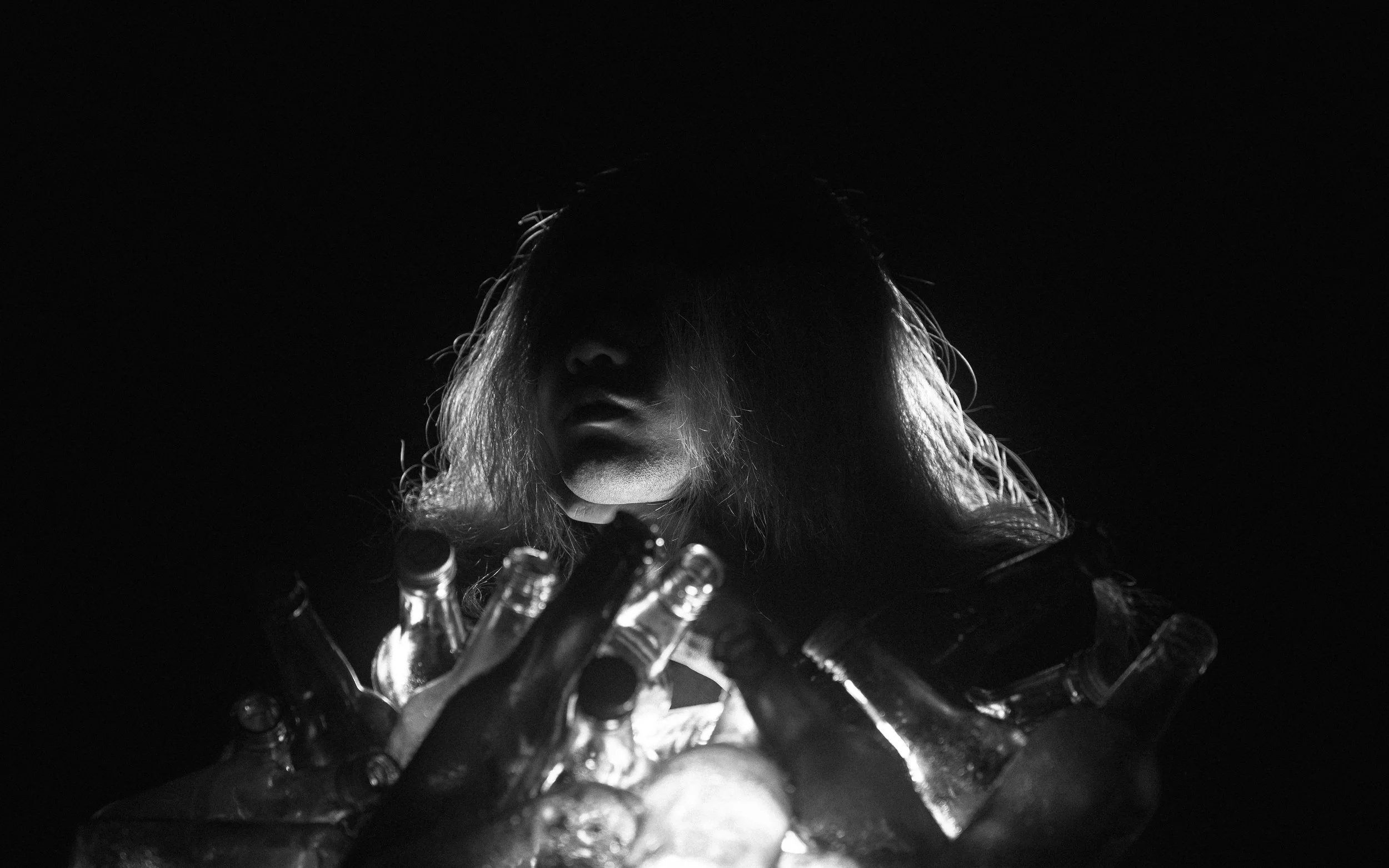 A person with long hair, lit from below, faces downward with their eyes closed, surrounded by several glass bottles.