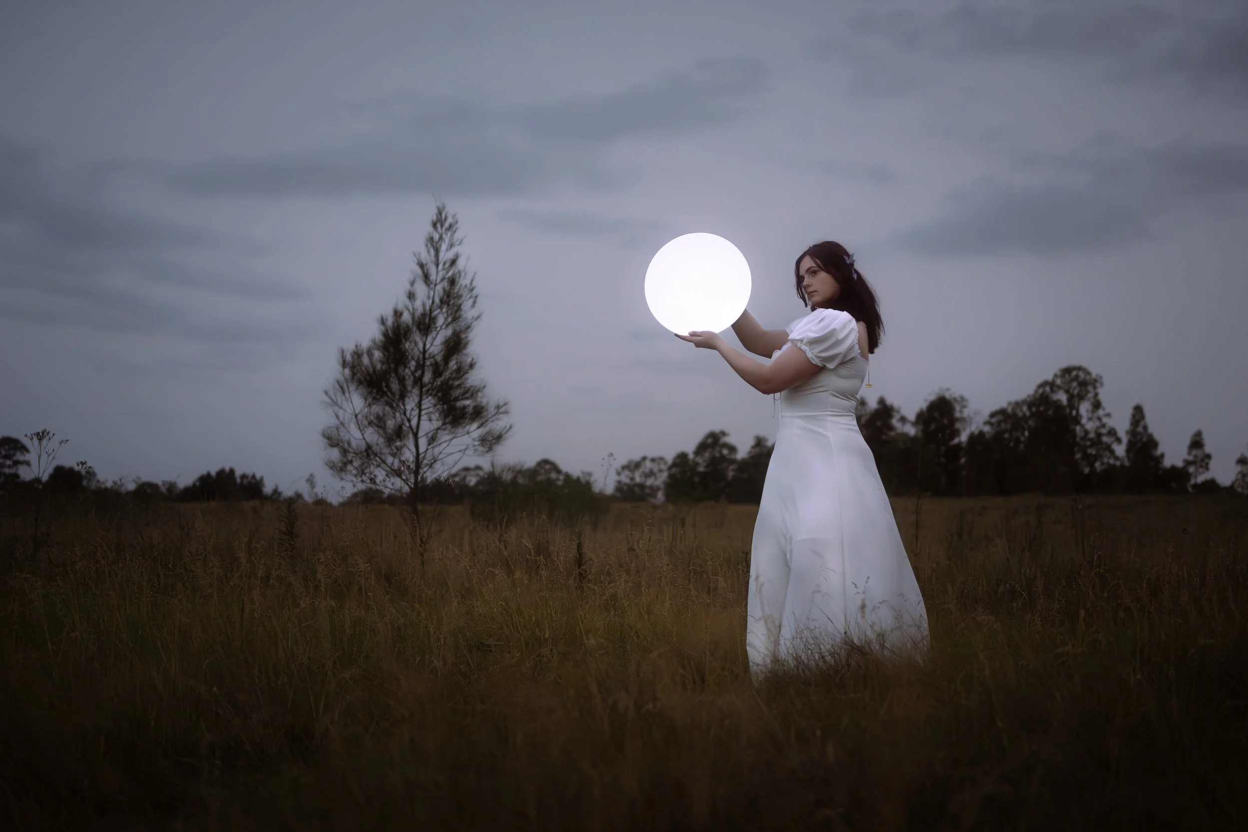 A woman in a white dress standing in an open field at dusk, holding a glowing orb that resembles a full moon. Dark clouds and trees are visible in the background.