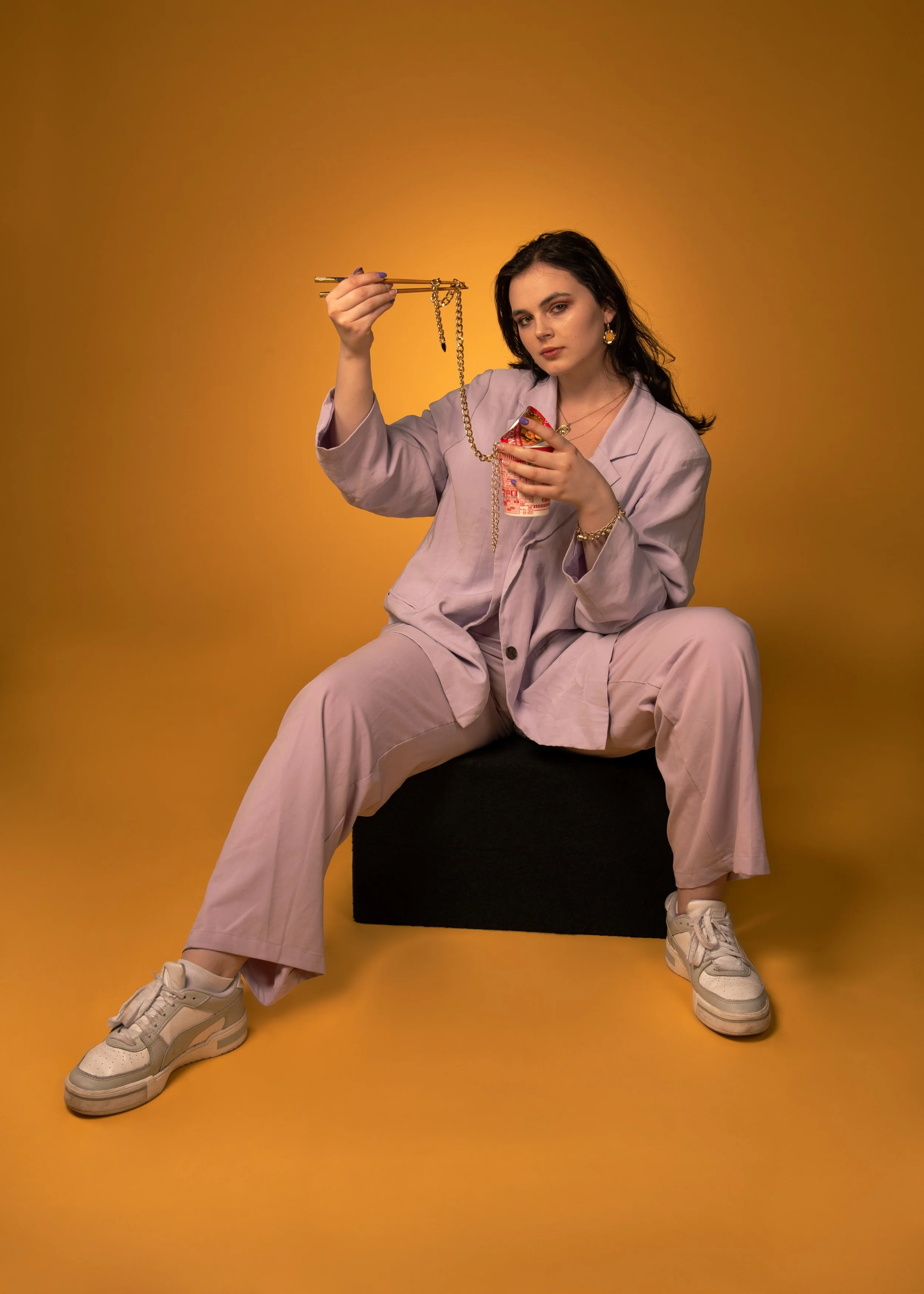 A young woman in pastel pink outfit sitting on a black cube against a yellow backdrop, holding chopsticks with a gold chain and a can of soda.