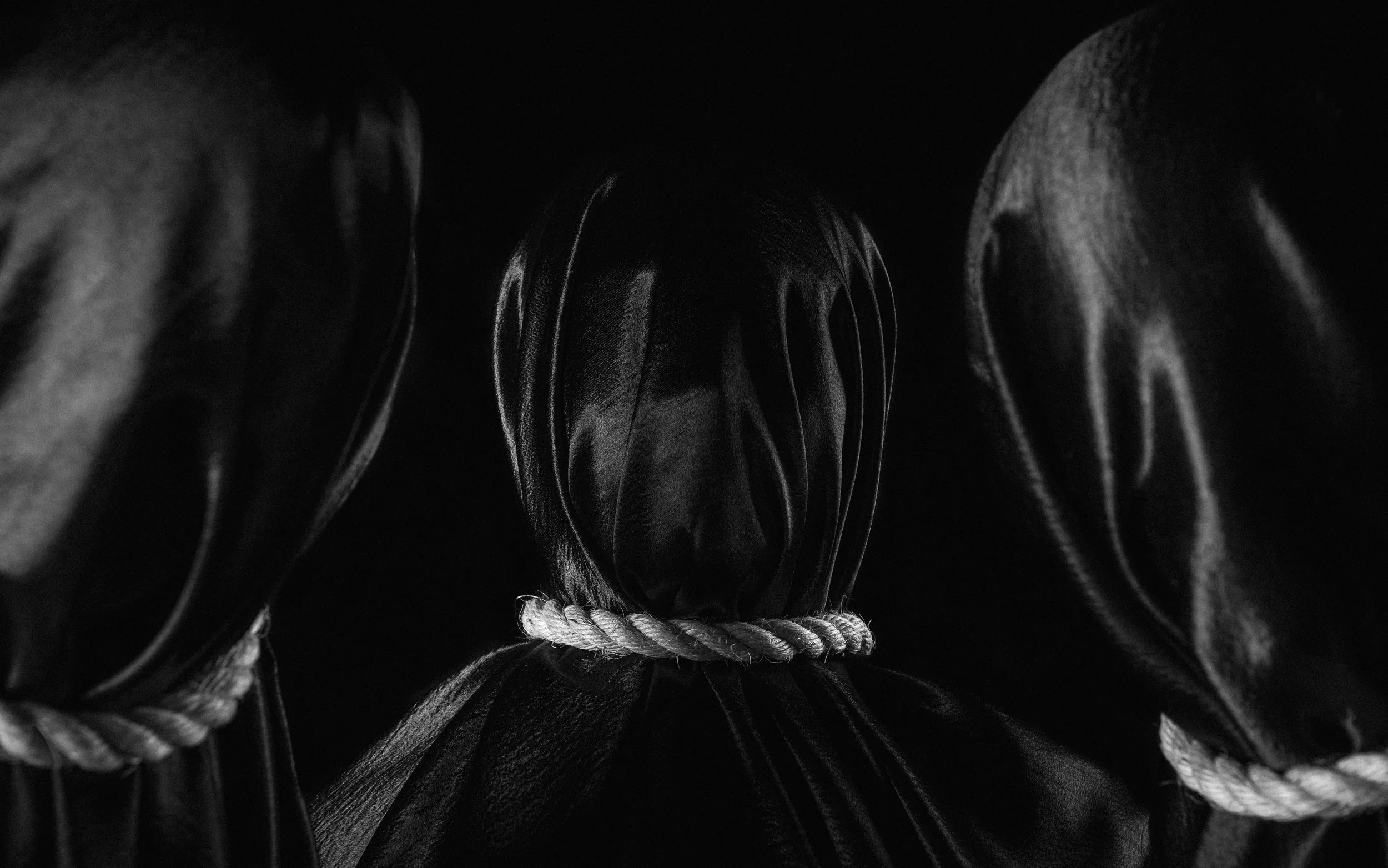 Black and white photo of a hooded figure with their head bowed, wearing a dark hoodie and a rope tied around their neck.