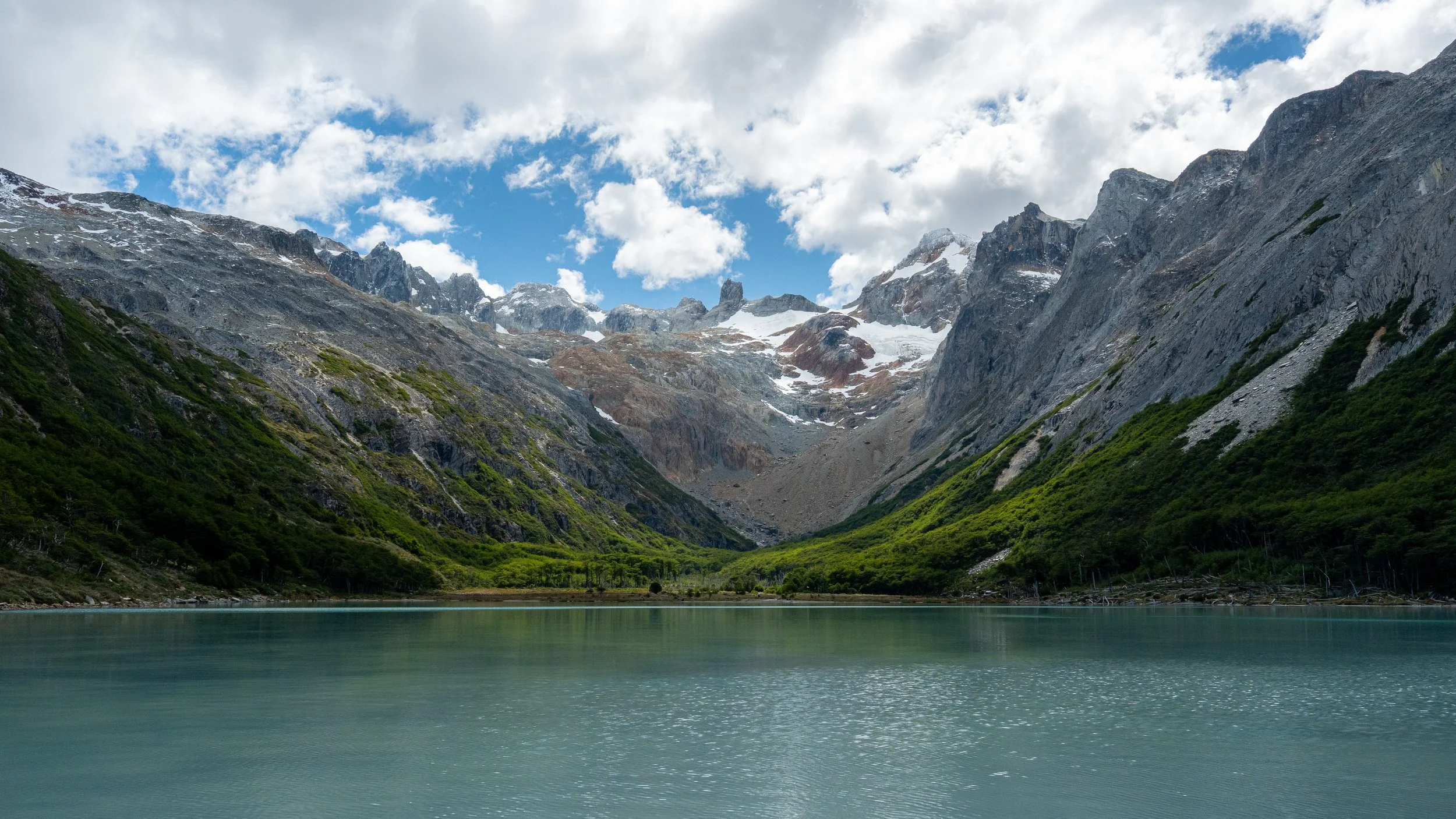 Scenic view of a mountain landscape with snow-capped peaks, green forested slopes, and a calm body of water in the foreground under a partly cloudy sky.