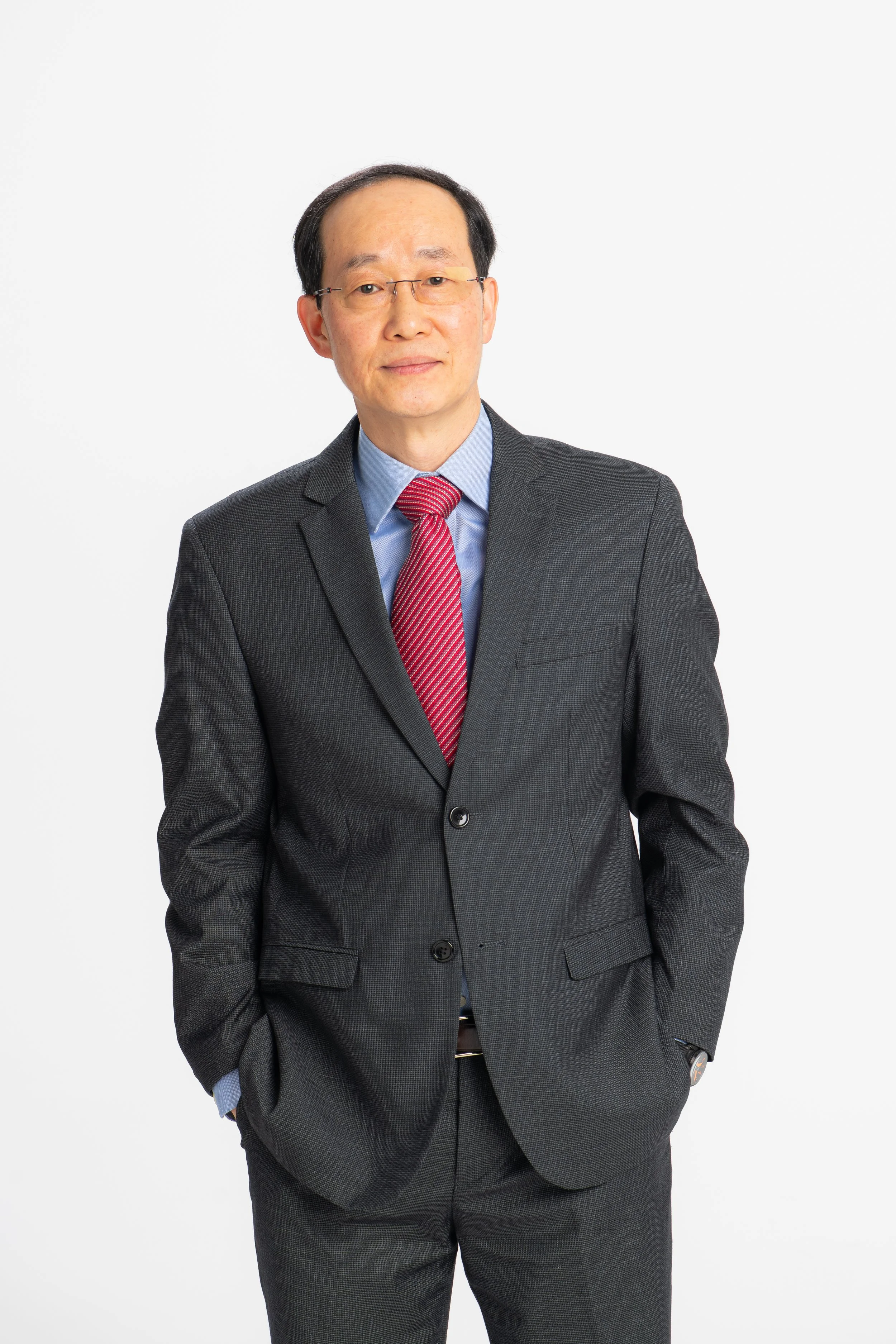 A man in a gray suit with a red striped tie and glasses, standing against a plain white background.