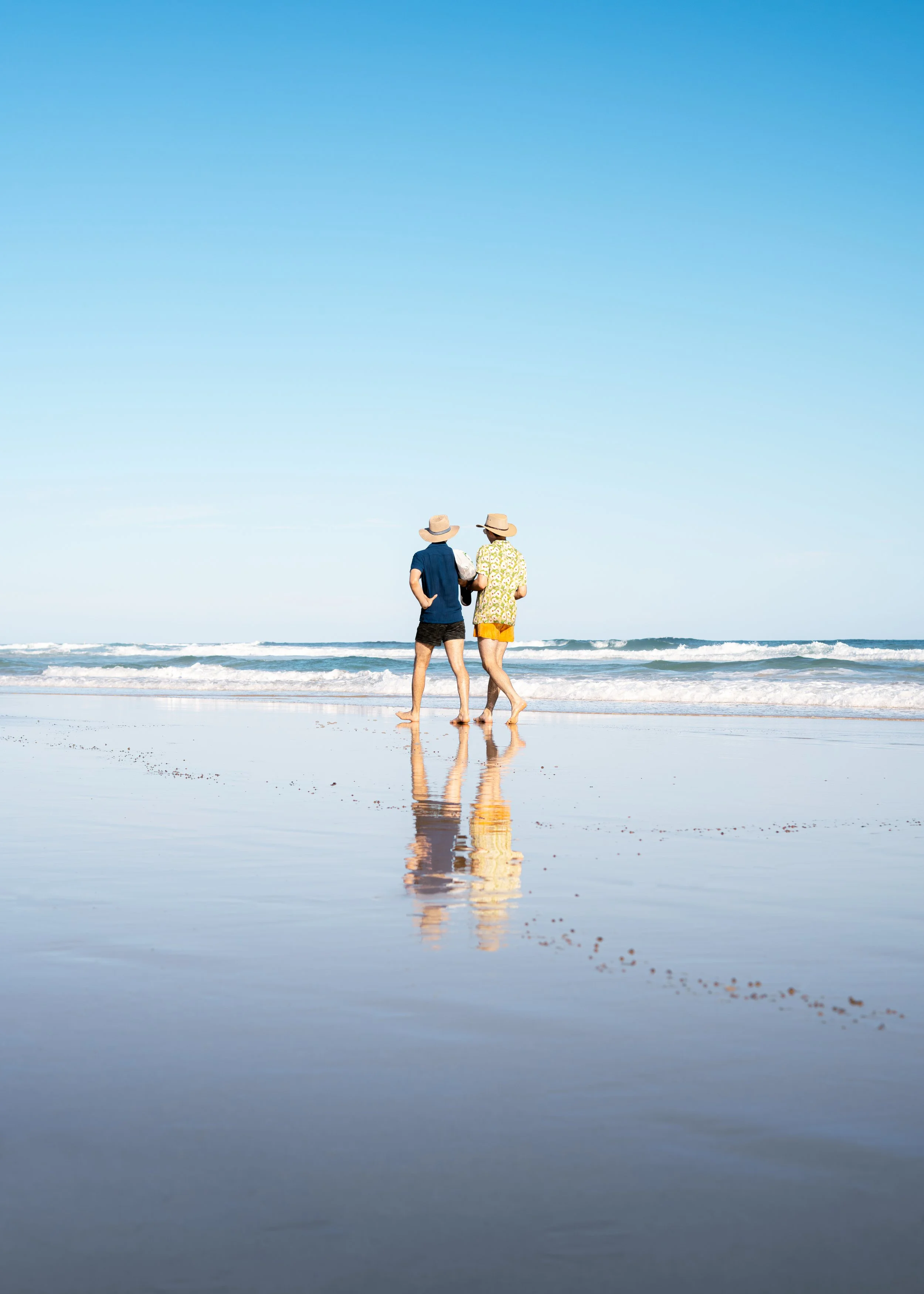 Two people walking on the beach, wearing hats, with the ocean waves in the background, and their reflection visible on wet sand.
