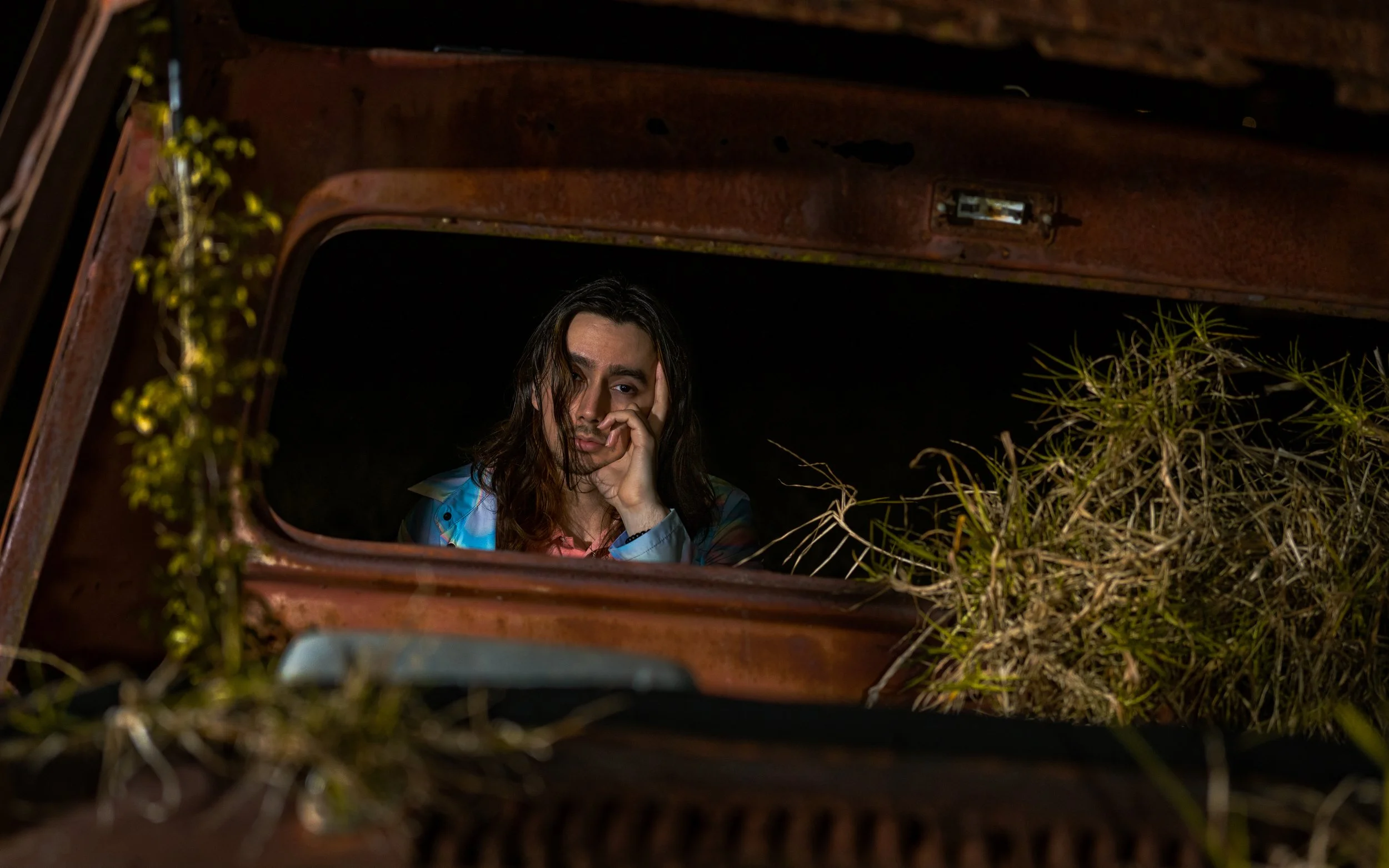 A person with long dark hair resting their face on their hand, looking through the window of an abandoned, rusty vehicle surrounded by overgrown plants at night.
