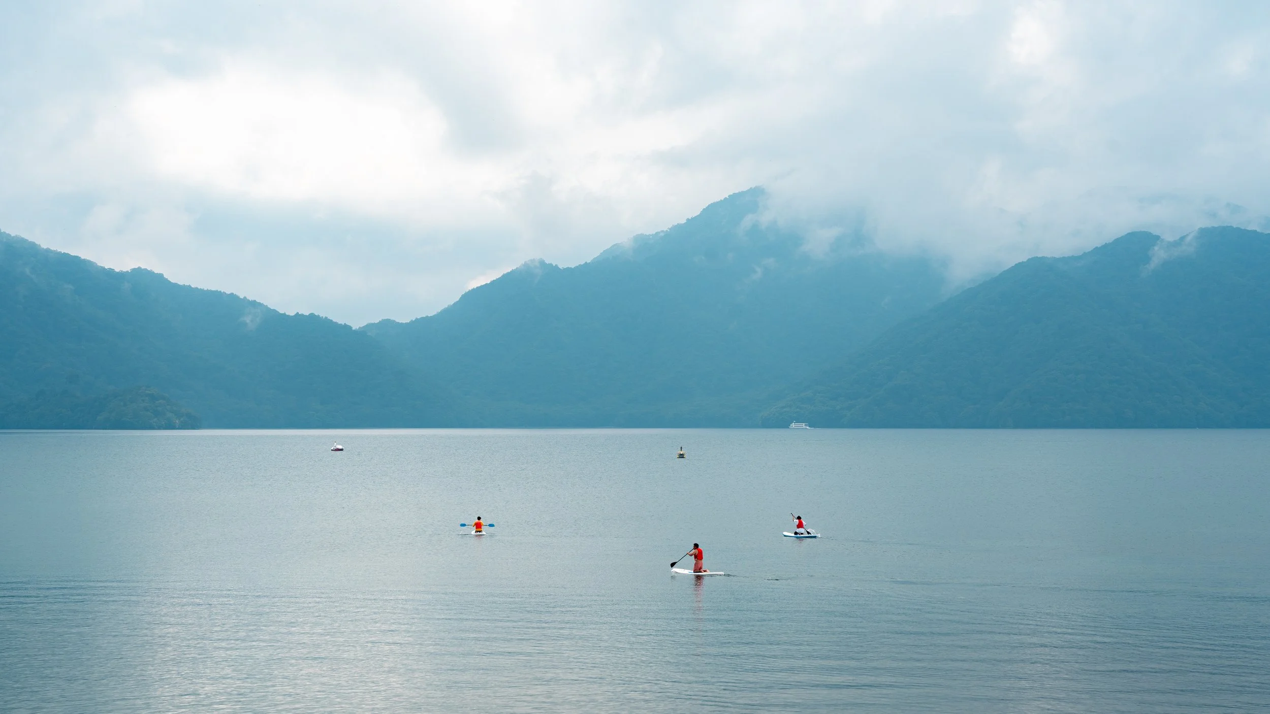 People kayaking on a calm lake with mountains and cloudy sky in the background.