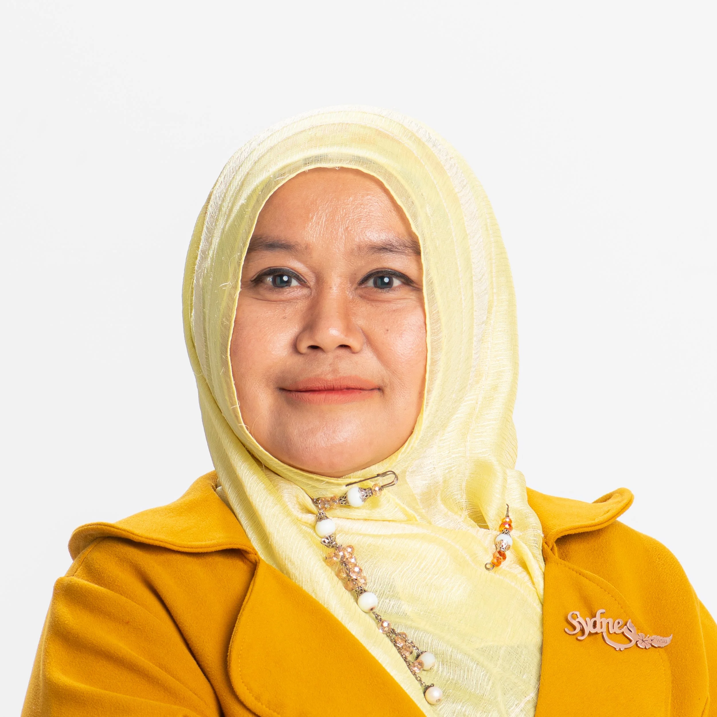 Close-up of a woman wearing a yellow hijab and a yellow jacket, smiling, with a jewelry necklace, against a plain white background.
