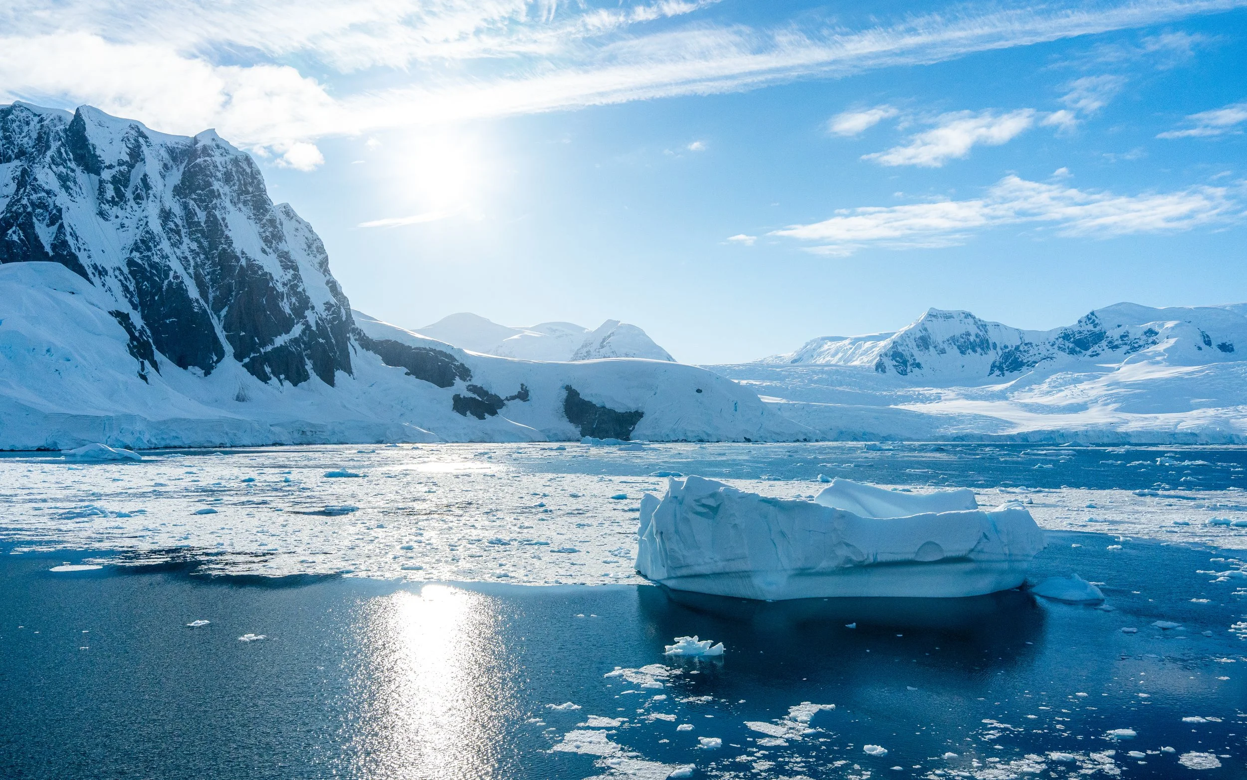 Snow-covered mountains and icy waters with floating ice chunks under a partly cloudy sky and bright sunlight.