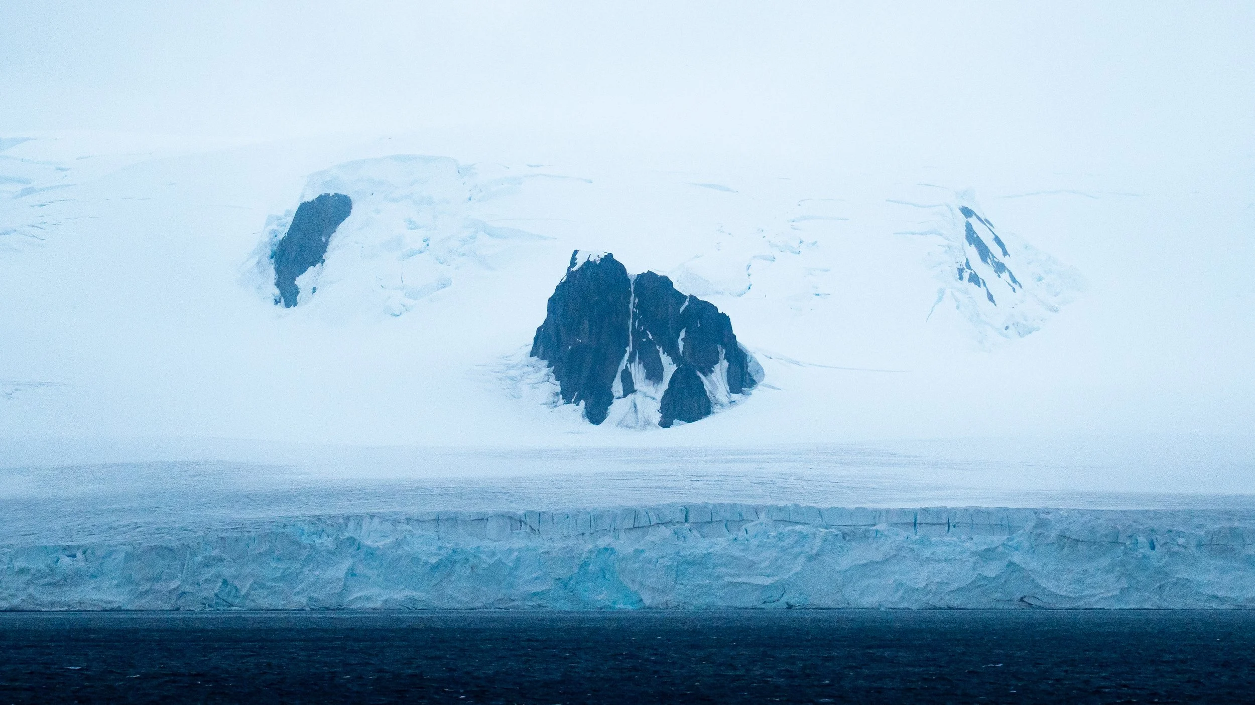 A polar landscape featuring a large snow-covered mountain with exposed dark rock faces, an icy body of water in the foreground, and a layer of ice along the edge of the water.