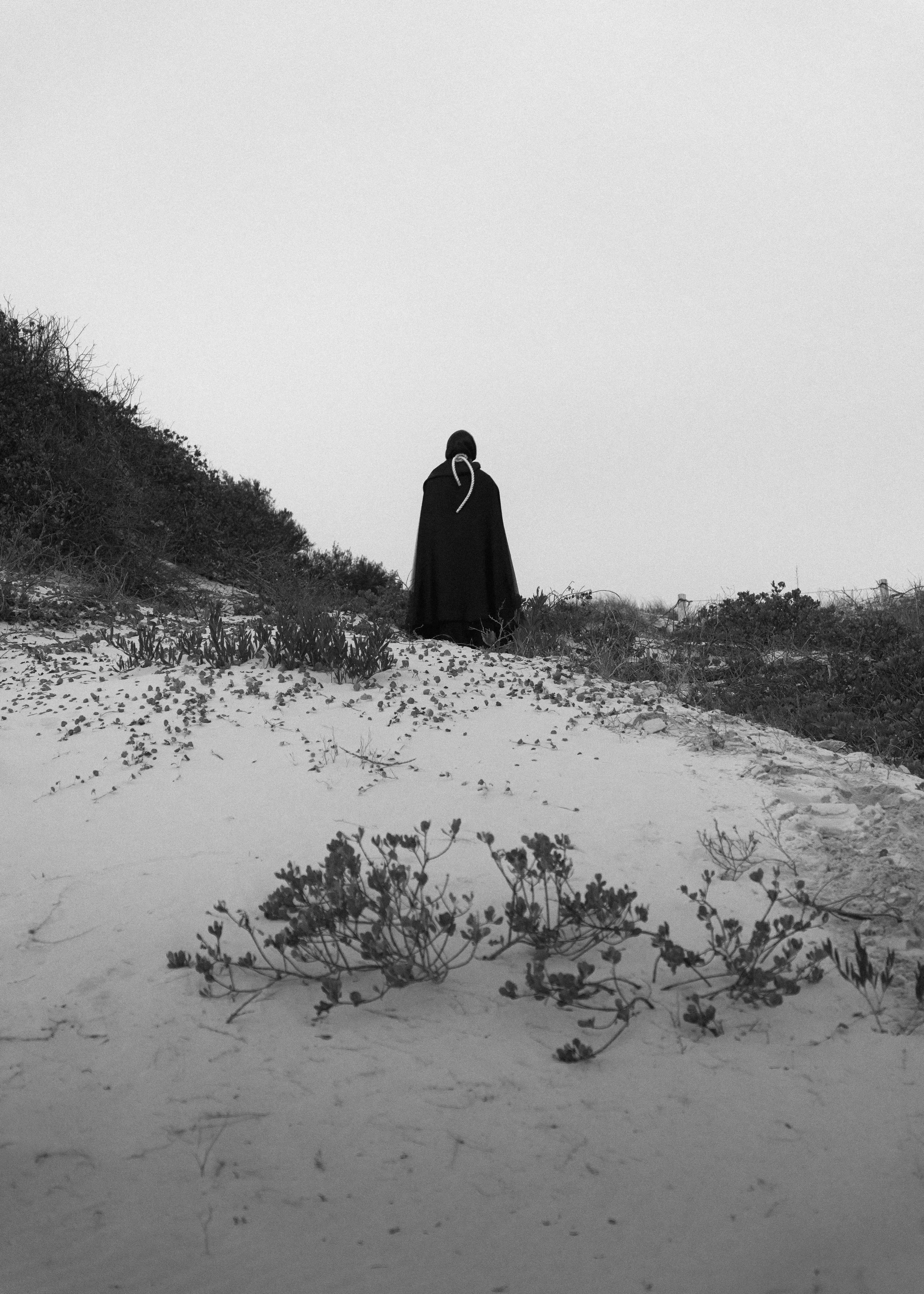 A person wearing dark clothing and a hood standing alone on a sandy beach with sparse vegetation, facing away from the camera, amidst a cloudy sky.