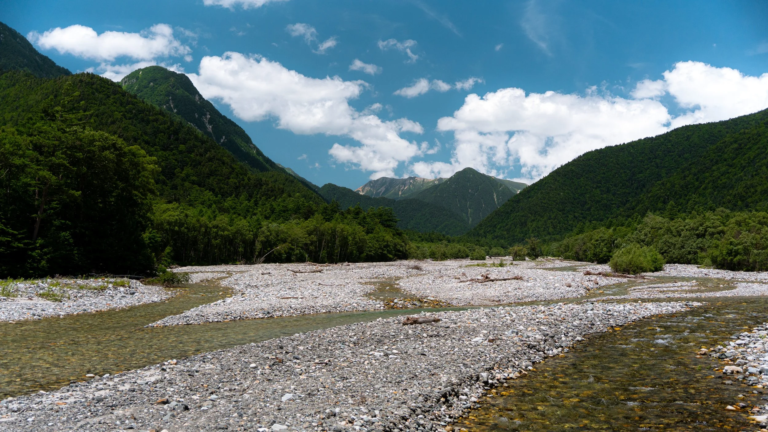 A mountain landscape with green forested slopes and a rocky riverbed under a partly cloudy blue sky.