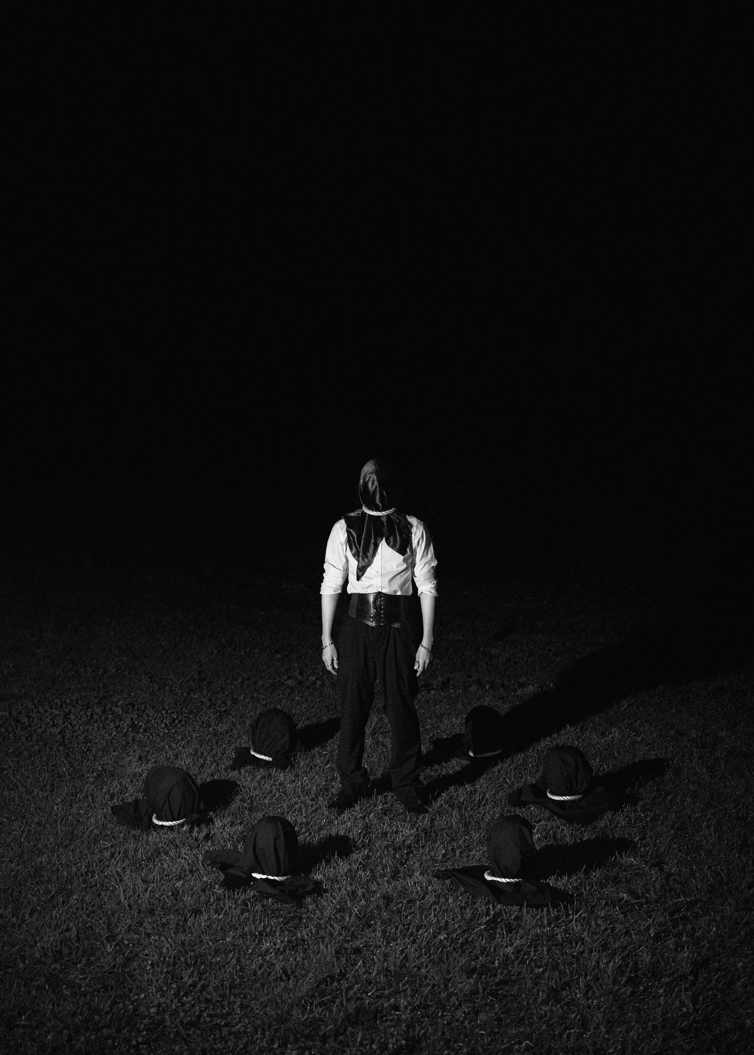 Person dressed in dark clothing, standing on grass at night, surrounded by a circle of black hoods with white strings.